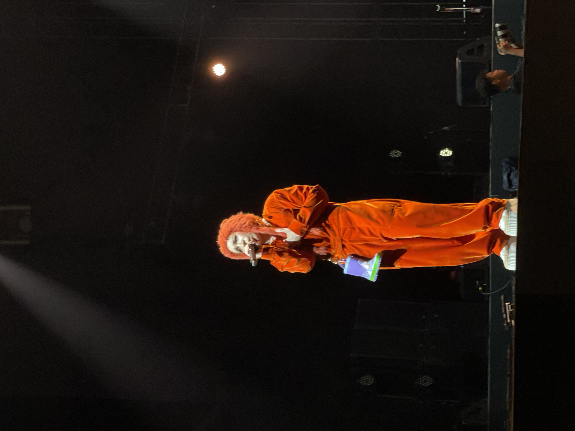 A person with a red afro wearing a red jumpsuit and white sneakers is performing on a dark stage with a microphone in their hand. The person is holding a purple and green object in their left hand. Stage lighting is visible. Another person with a camera is visible on the right side of the image. It is not possible to determine the location in Salembaran, Indonesia from this image.