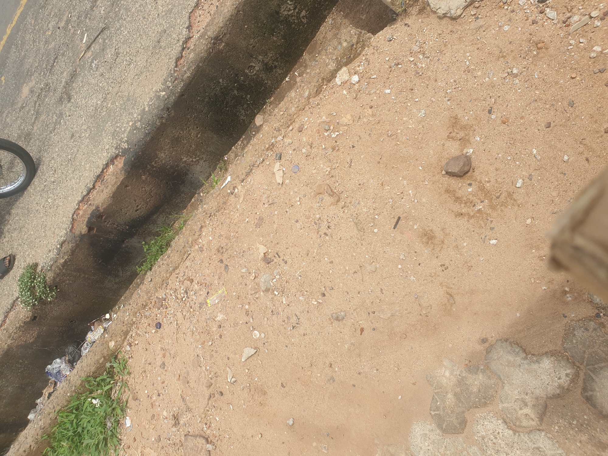 The image depicts a roadside scene in Adankolo, Nigeria. A concrete drainage ditch, filled with stagnant water, trash, and some green vegetation, runs parallel to an asphalt road. The road edge is broken and uneven. On the other side of the ditch, there is a dirt/sand area mixed with gravel and litter. Part of a bicycle wheel and a foot are visible near the road. A section of patterned concrete paving stones can be seen in the lower right corner, along with a blurry image of part of a post, possibly a wooden support. The environment appears unkempt with some natural growth interspersed with human refuse.