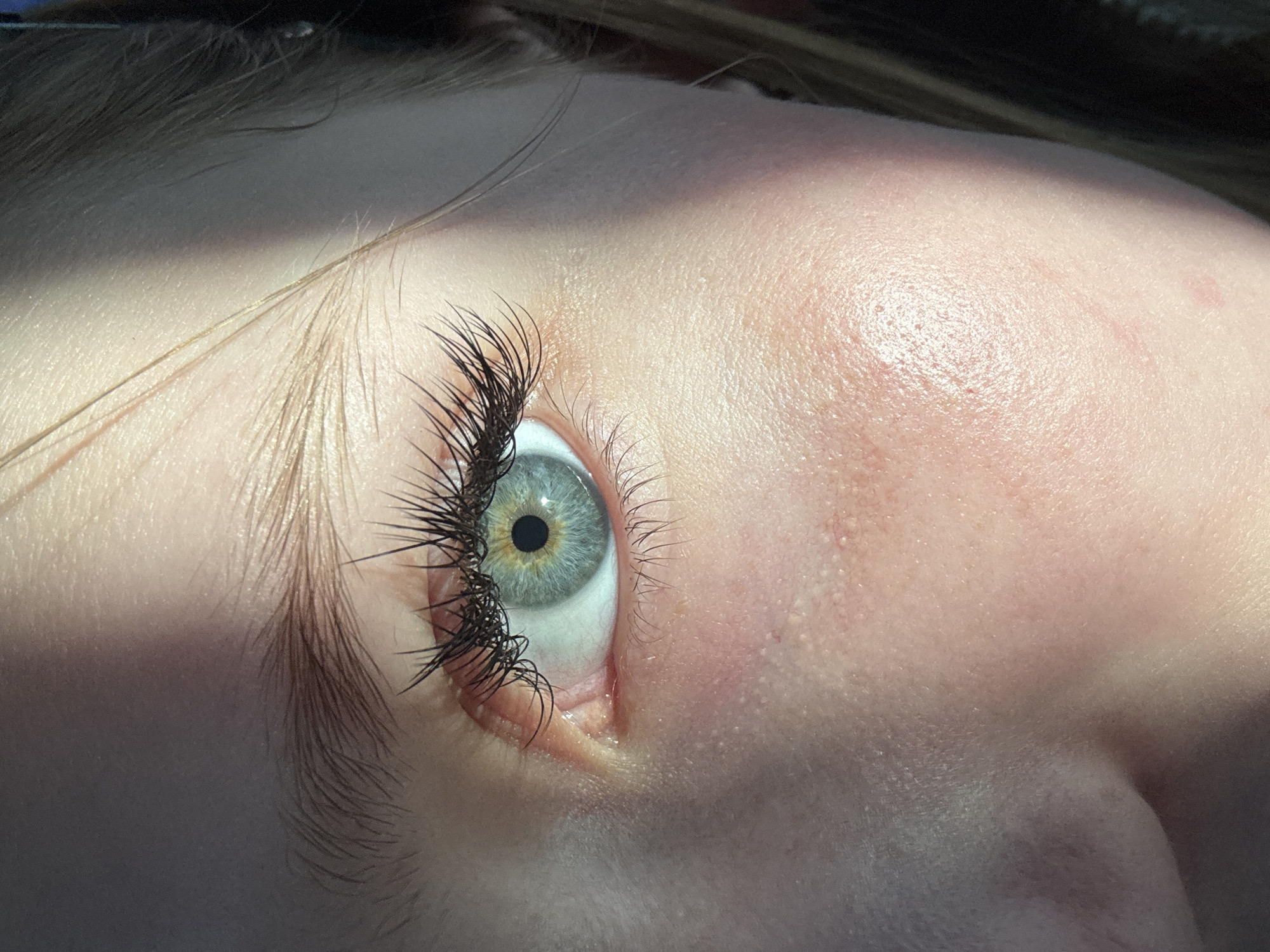 A close-up photograph shows a human eye with long, dark eyelashes. The eye's iris is a mix of blue and green shades, with a dark pupil in the center. The surrounding skin is light-colored and shows some texture and minor imperfections. Light reflects off the skin and eye, creating highlights and shadows. The subject's face is angled towards the top of the frame. The location of the image is not determinable from the image alone. Berezniki, Russia, cannot be determined from the image alone.