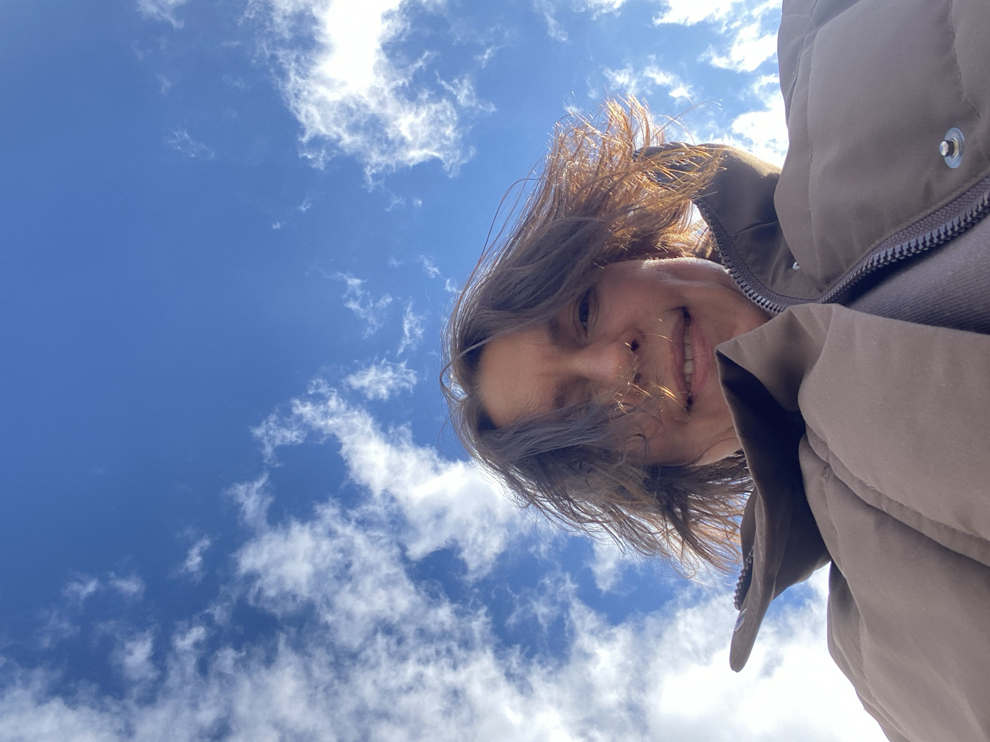 A low-angle, eye-level shot shows a person with short, light brown hair and a brown jacket, taken against a partly cloudy blue sky. The individual is smiling slightly. The setting does not appear to be Nizhnie Yaki, Russia.