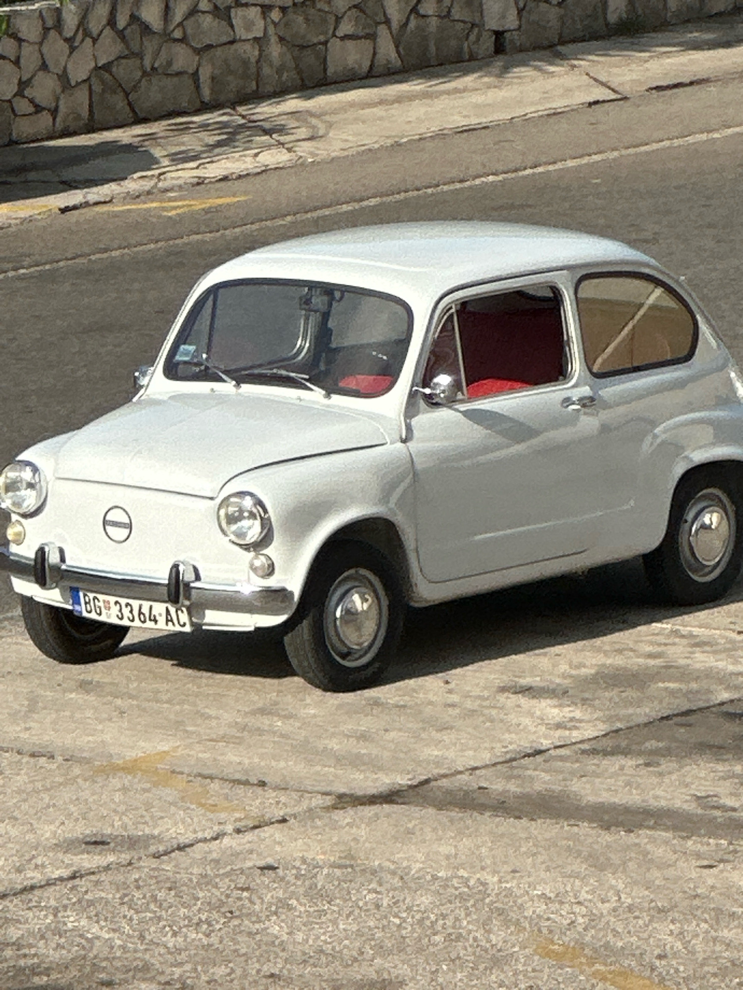 A white vintage Zastava 600, also known as a Fića, is parked on a concrete surface in Budva, Montenegro. The car is facing to the left, with its front slightly angled towards the camera. The license plate reads "BG 3364-AC". Behind the car, a stone wall lines the upper part of the image, suggesting a sloped terrain. The ground is a textured concrete with some faint yellow markings. The overall lighting indicates a sunny day, casting shadows. There are no people visible in the frame. The image captures a static scene, focusing on the classic automobile.