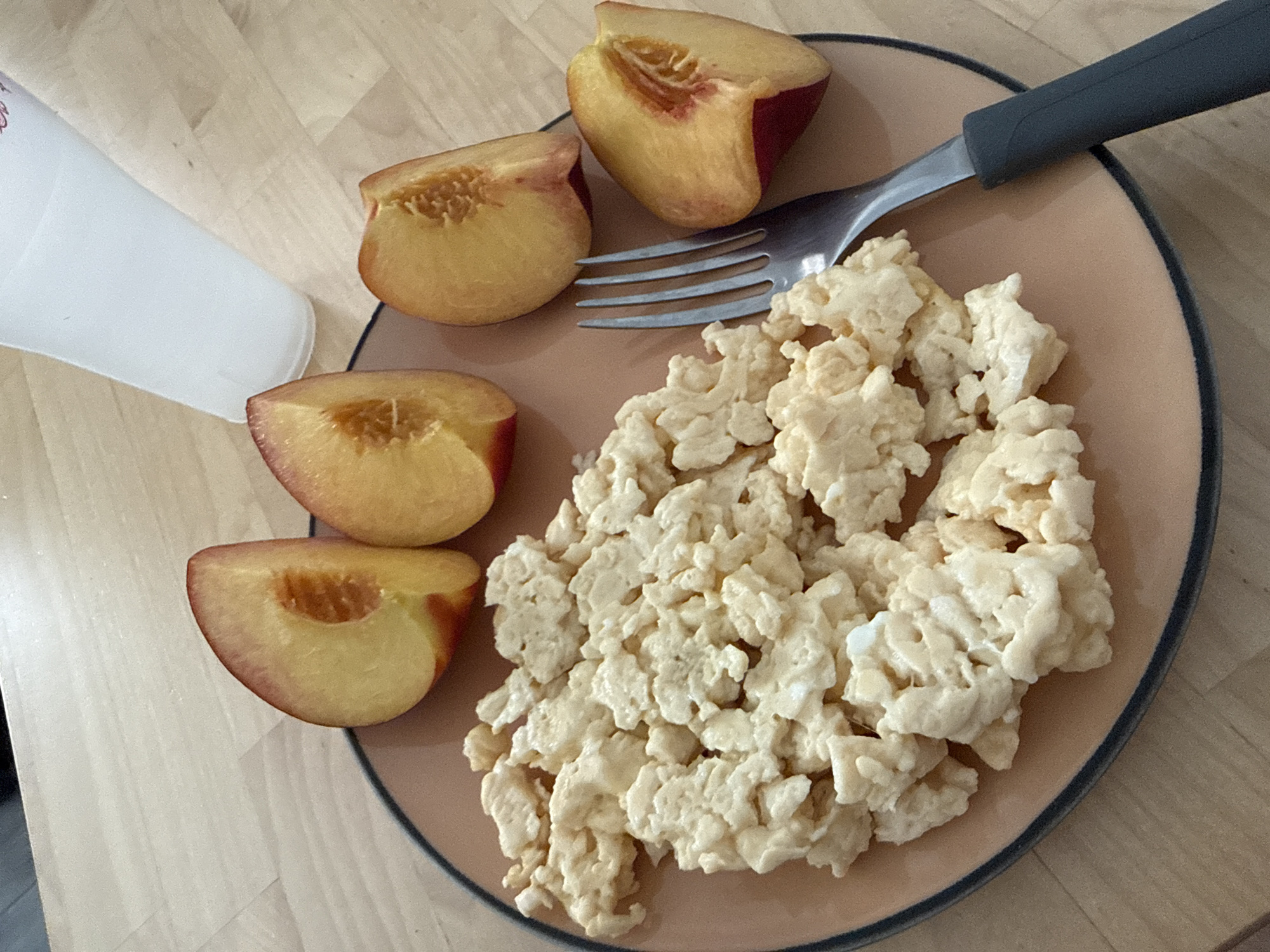 A close-up, overhead shot shows a plate of scrambled eggs and sliced peaches on a wooden table. A white plastic cup is in the upper right corner. The plate is tan with a dark blue rim and contains a generous portion of fluffy, pale yellow scrambled eggs. Four slices of peaches are arranged around the eggs. The peaches are a vibrant yellow with reddish-pink skin and show their seed cavities. A silver fork with a dark gray handle rests on the right side of the plate, partially submerged in the scrambled eggs. The wooden table has a light, natural grain. The scene appears to be an indoor setting, possibly a home or cafe, and there is no specific information to indicate its location in Almada, Portugal.
