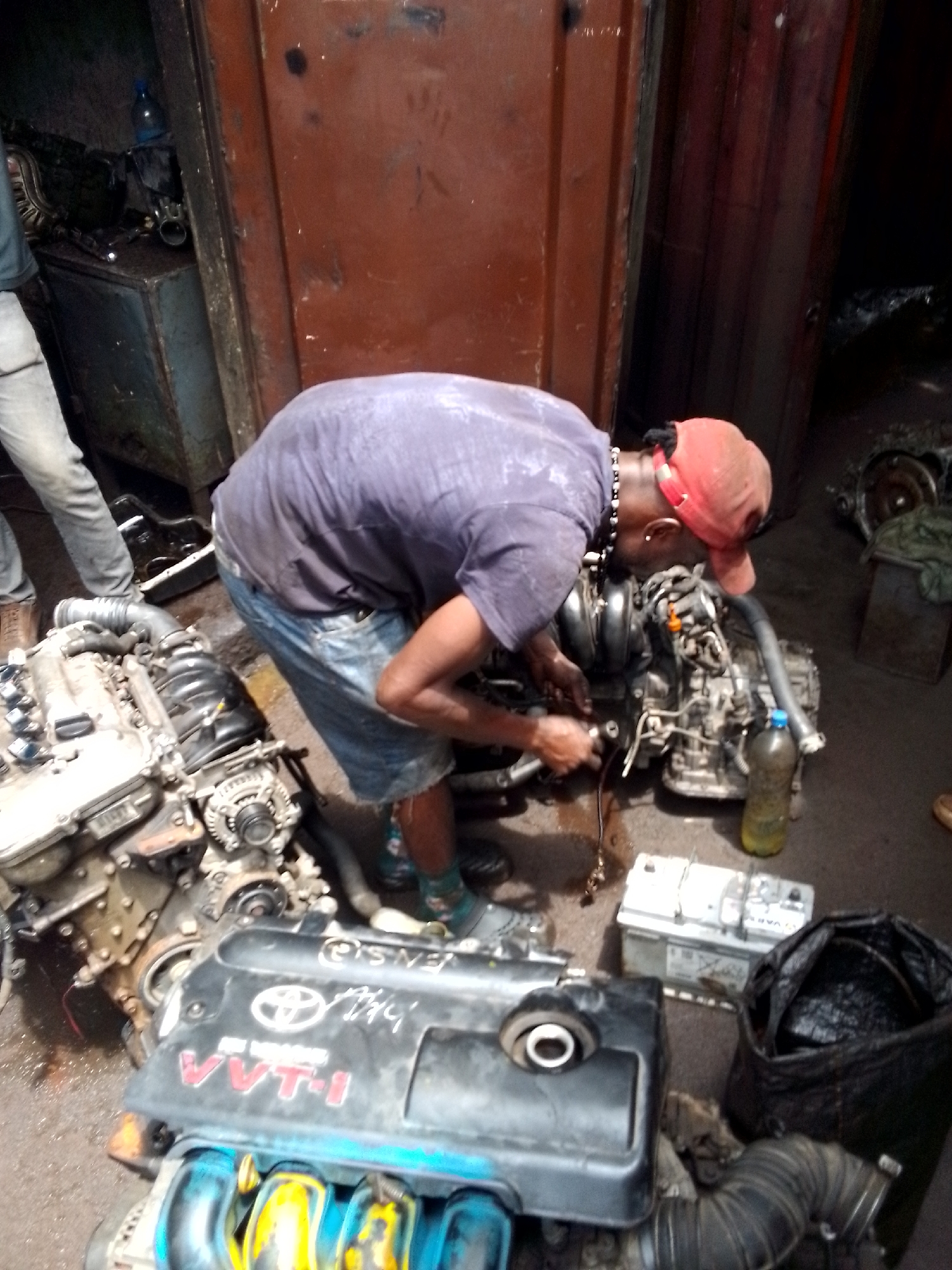 In Onitsha, Nigeria, an auto mechanic wearing a purple t-shirt, jean shorts, red cap, and a beaded necklace works on a car engine. The mechanic is bending over an engine on the ground. Several other engines and parts, including a black engine cover labeled "VVT-I" and a car battery, are scattered around the workspace. A man in grey pants stands nearby. The environment appears to be a small, cluttered garage.