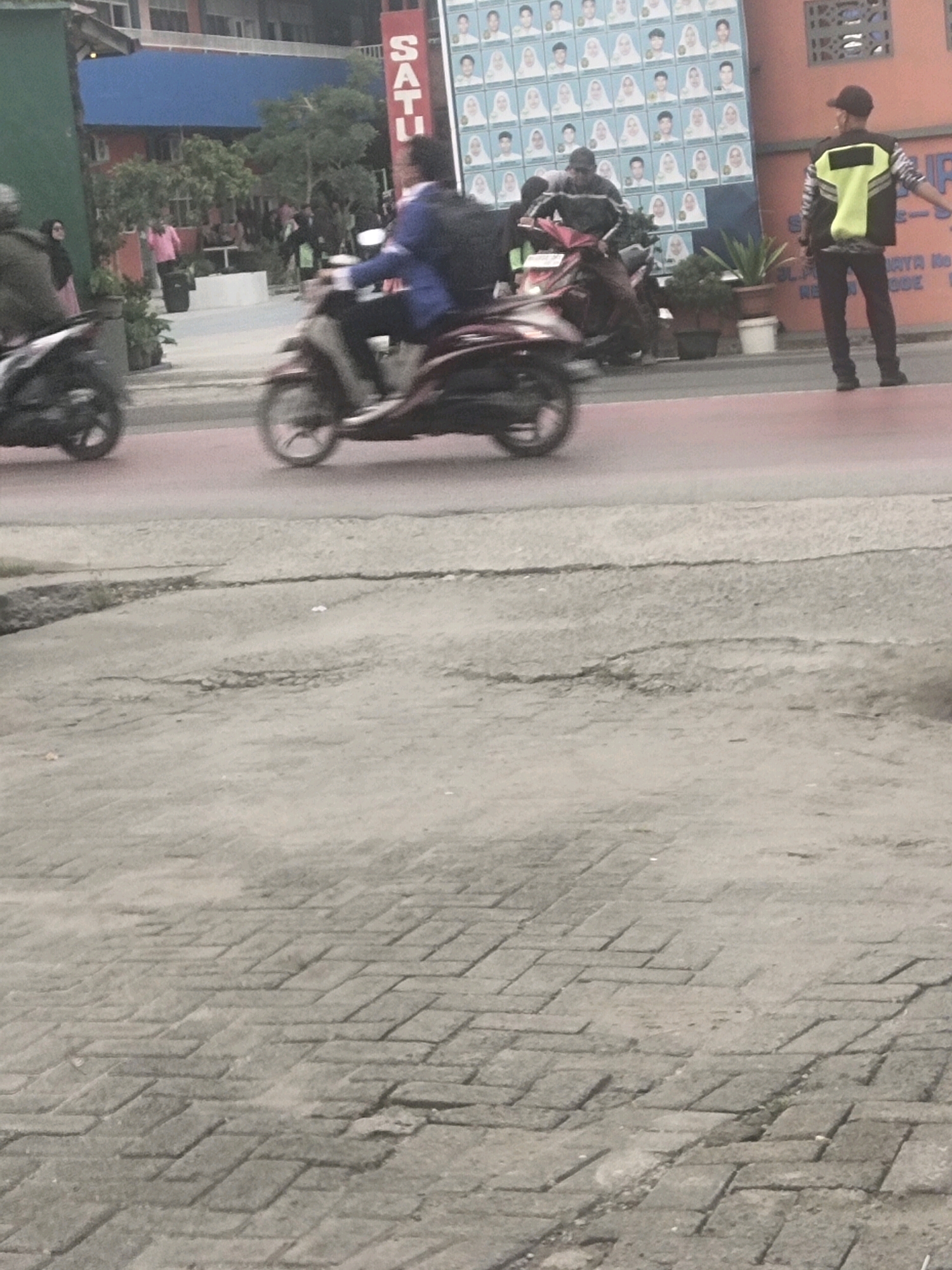 The image depicts a street scene in Medan, Indonesia. In the foreground, a paved area with a brick pattern is visible. The middle ground shows a paved street with two individuals on motorcycles in motion. The motorcycle on the left has its rear wheel and part of its frame visible, while the motorcycle on the right is more clearly seen with a rider wearing a blue jacket and backpack. To the right of the street, a person in a yellow safety vest and a dark cap is standing, appearing to be directing traffic or observing the scene. Behind them, a large banner with a grid of student portraits is displayed on a building. A red building with a blue roof is also visible, along with trees and some potted plants. The overall impression is a busy urban environment.