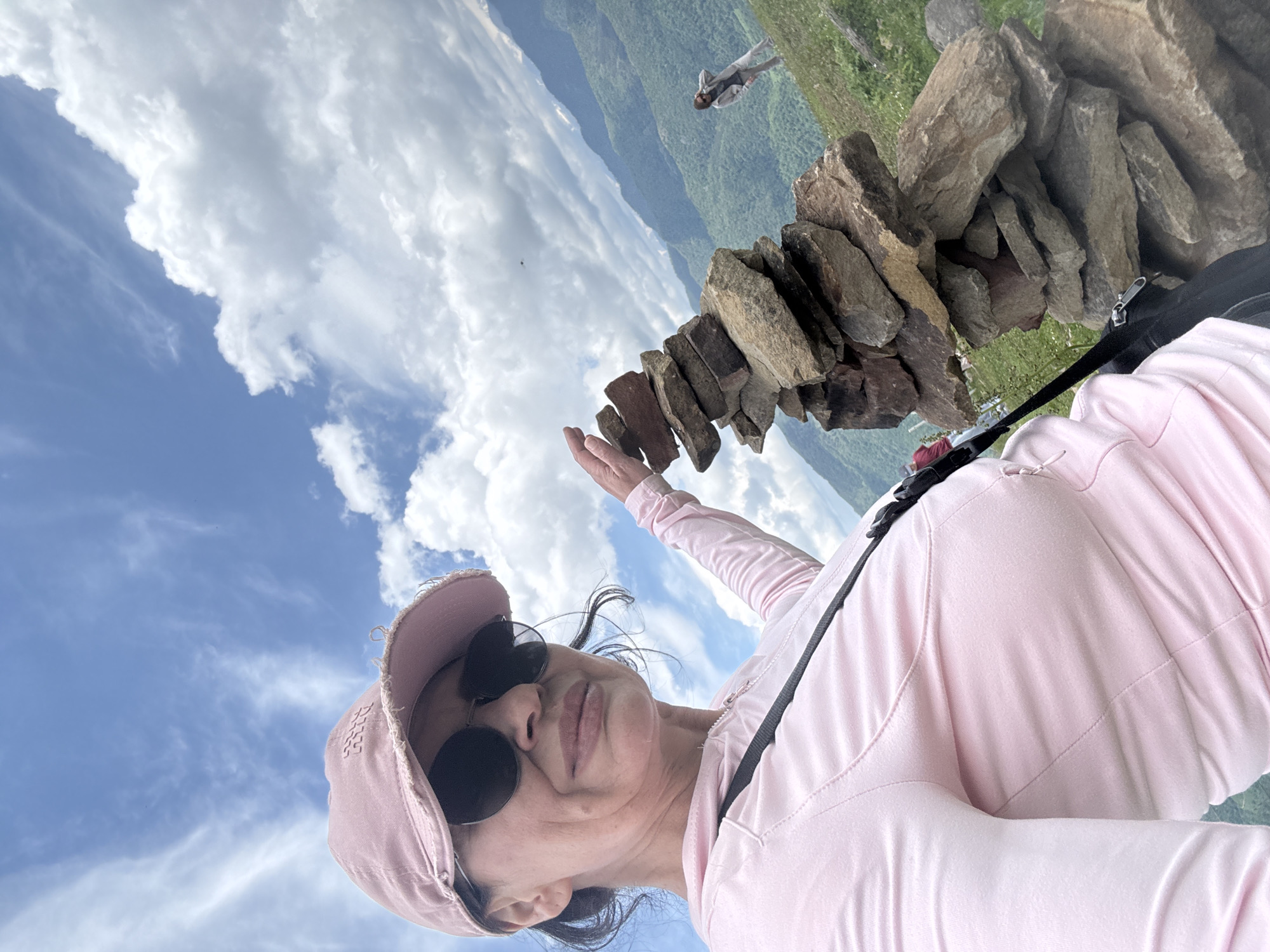 In an outdoor, elevated landscape scene likely in the vicinity of Nizhnyaya Teberda, Russia, a woman wearing a pink baseball cap and sunglasses is taking a selfie. She is positioned to the left of the frame, her face partially obscured by the cap. Her right hand is extended towards a stacked cairn of stones located to her right. She is wearing a pink long-sleeved shirt. The background reveals a vast expanse of rolling green hills under a sky filled with white, billowy clouds. In the distance, on a hillside, another person is visible, appearing to be walking. The overall environment suggests a mountainous or high-altitude terrain.