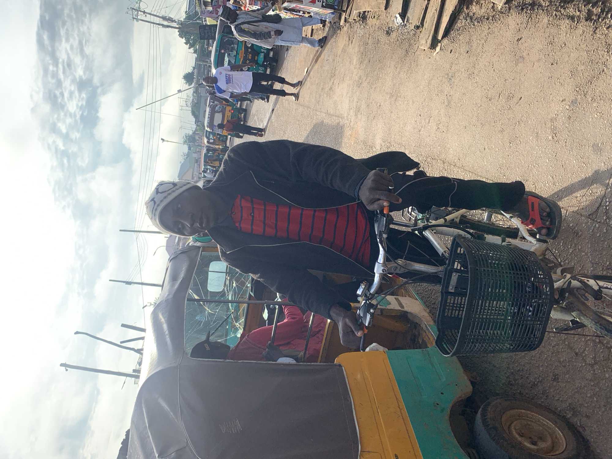 A Black man wearing a striped red and black shirt, a black jacket, black pants, red sandals, and a white beanie with a pattern is riding a bicycle on a dusty road. He is in the foreground and is the main subject of the image. To his left is a three-wheeled vehicle, likely a tuk-tuk, with a dark canopy and yellow and green body. The tuk-tuk's windshield appears cracked. In the background, there are more tuk-tuks and several other people walking. The scene appears to be a busy street in Jos, Nigeria, given the presence of tuk-tuks which are common modes of transportation there. The sky is partly cloudy with a bright sun, suggesting it is daytime. The road is unpaved and appears to be made of dirt and gravel. There are utility poles and wires visible in the background, indicating an urban or semi-urban setting.