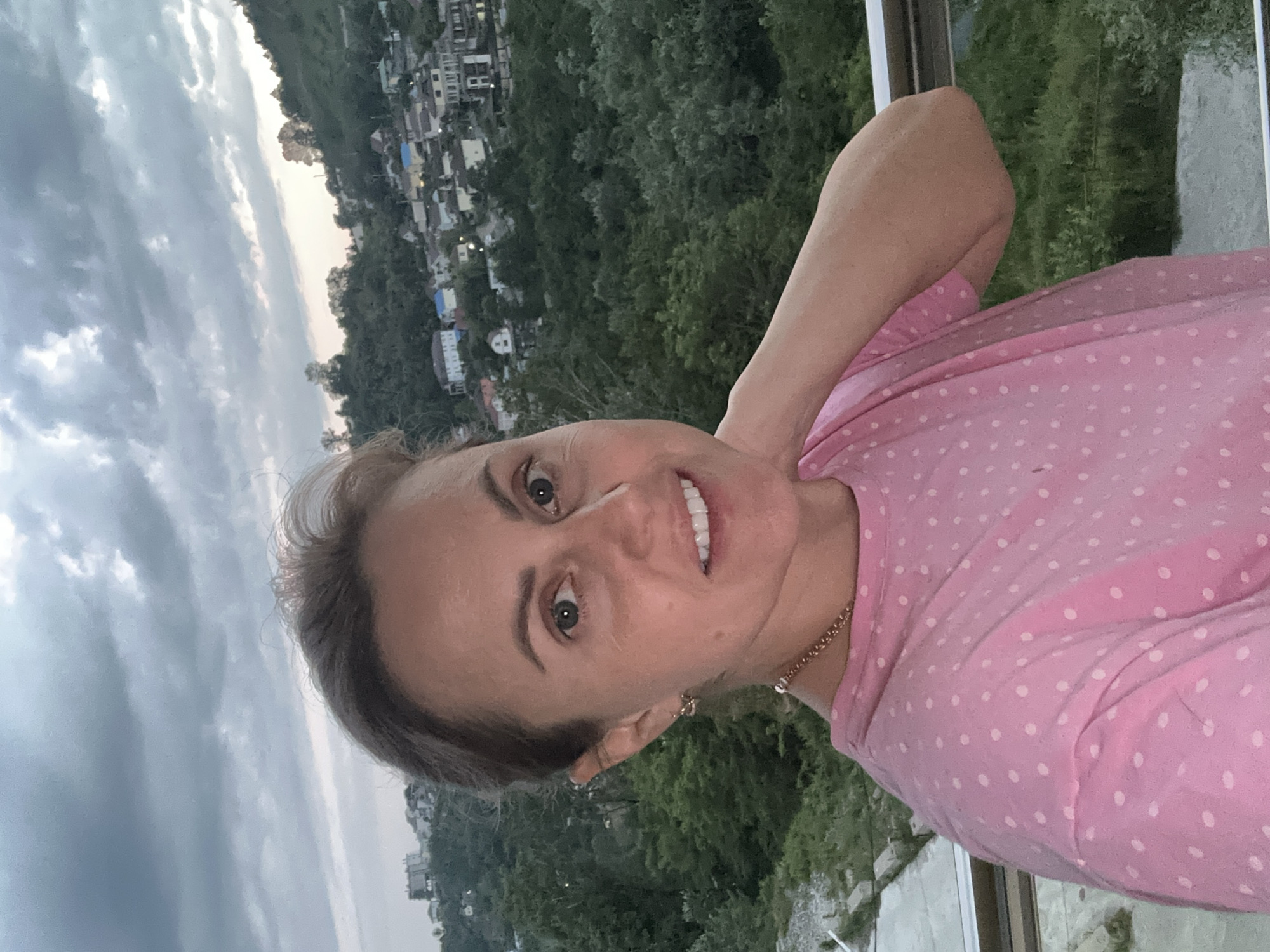 A medium shot captured outdoors shows a woman with light skin and brown hair pulled back. She is wearing a pink t-shirt with white polka dots and a gold necklace. Her right arm is bent and resting on her neck, and her left hand is not visible. She is smiling and looking directly at the camera. The background features a hillside dotted with houses, lush green trees, and a cloudy sky. The scene is likely located in the city of Razdolnoye, Russia, a small town known for its hilly terrain and scattered residential areas.