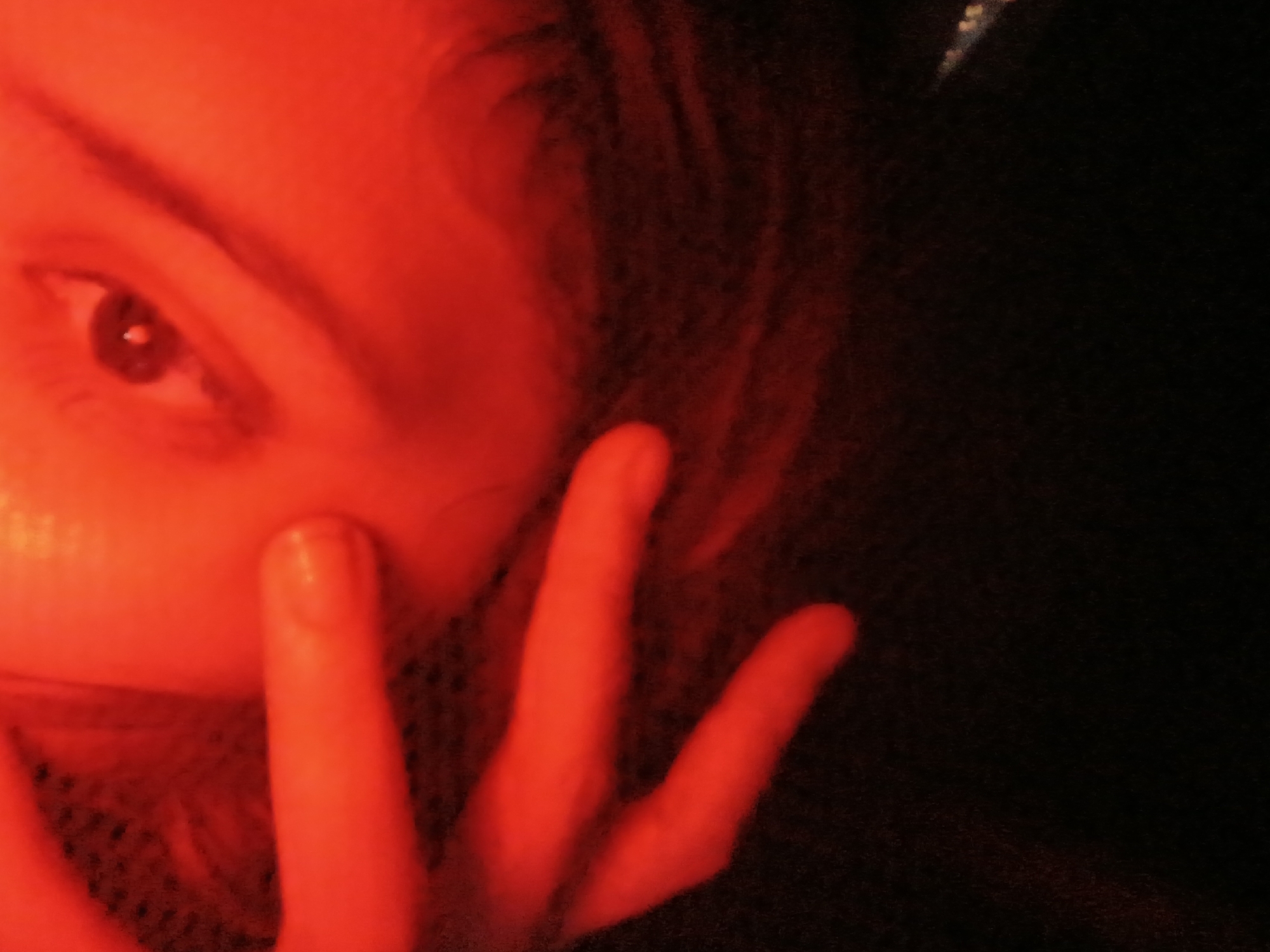 A close-up photograph, bathed in a pervasive red light, focuses on the face and hand of an individual. The face, partially obscured, reveals an eye with a visible iris and eyelashes. The hand, with extended fingers, is positioned near the face. The background is dark and indistinct. The photograph's red hue obscures details and makes identification difficult. Location: This image is not indicative of a specific location in Buenos Aires, Argentina, or any other city. It is a close-up studio shot with artificial lighting.