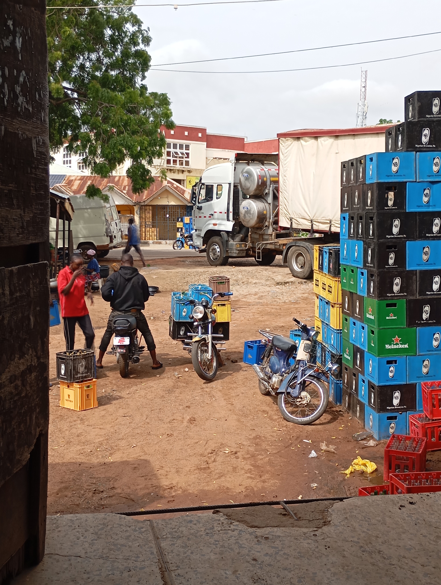 In Shendam, Nigeria, on a dirt ground surface, a scene unfolds with various vehicles and stacked crates. In the foreground, a person wearing a black hoodie is seated on a motorcycle, facing away from the camera. To their left, a person in a red shirt and black pants stands, gesturing with their hands. In front of the person in red, there are two black crates stacked on top of two yellow crates. A blue motorcycle with a yellow basket and blue crates is parked on the right side of the frame. Behind the motorcycles, a large white truck with a covered trailer is parked, and to its left, a smaller white vehicle with its back door open. Several stacks of blue and black crates are visible, some displaying "NB Nigerian Breweries Plc." A few red crates filled with bottles are also present. In the background, buildings with reddish-brown roofs and white walls are visible, along with trees and a cell tower in the distance. The sky is overcast.