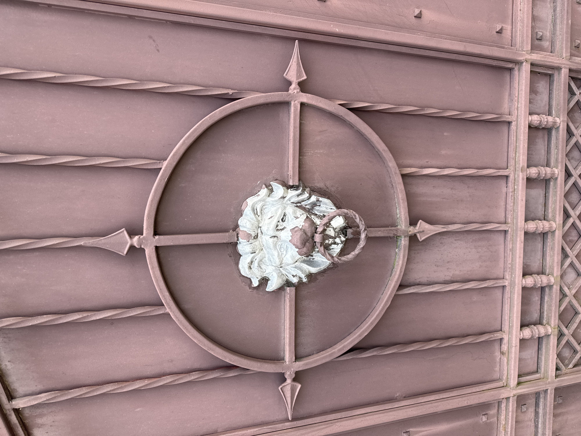 The image displays a weathered, reddish-brown metal gate or large door panel, located in the city of Odesa, Ukraine. The surface exhibits significant scuffing, fading, and discoloration, indicative of prolonged outdoor exposure.
At the center of the panel, a circular metal frame is intersected by vertical and horizontal metal bars, forming a prominent cross. The ends of these intersecting bars feature pointed, arrow-like finials.
Mounted centrally within this design is a lion's head door knocker. The lion's head is predominantly white or silver-toned, but substantial areas of paint have chipped and peeled, revealing the underlying reddish-brown metal, particularly on the mane, forehead, and muzzle. The lion grasps a simple, circular metal ring, matching the gate's reddish-brown hue, in its mouth, functioning as the knocker.
The gate structure includes multiple vertical, twisted metal bars positioned on either side of the central motif. Below the main lion's head assembly, a horizontal metal bar is visible, supported by a series of small, ornate, baluster-like elements. The lowest visible section of the gate features a lattice pattern composed of crisscrossing diagonal metal bars, forming a grid of diamond shapes. Square-headed rivets or bolts are visible along various structural joints and frames within the gate. No people or actions are depicted; the scene is static.