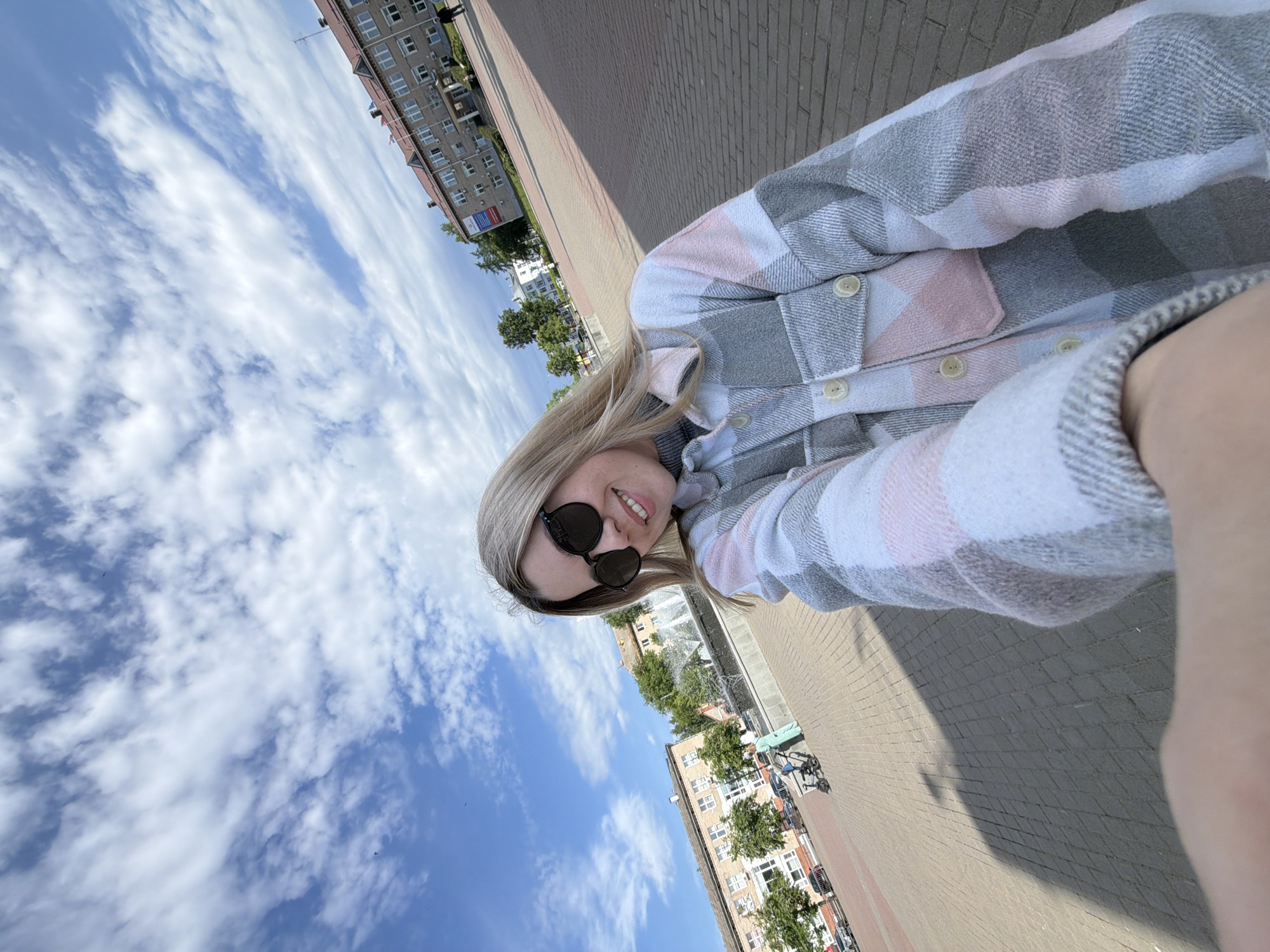 The image is a selfie taken by a woman wearing sunglasses and a plaid shirt. In the background, a building complex is visible, including a water fountain and a brick-paved ground. The location is Chaikovsky, Russia.