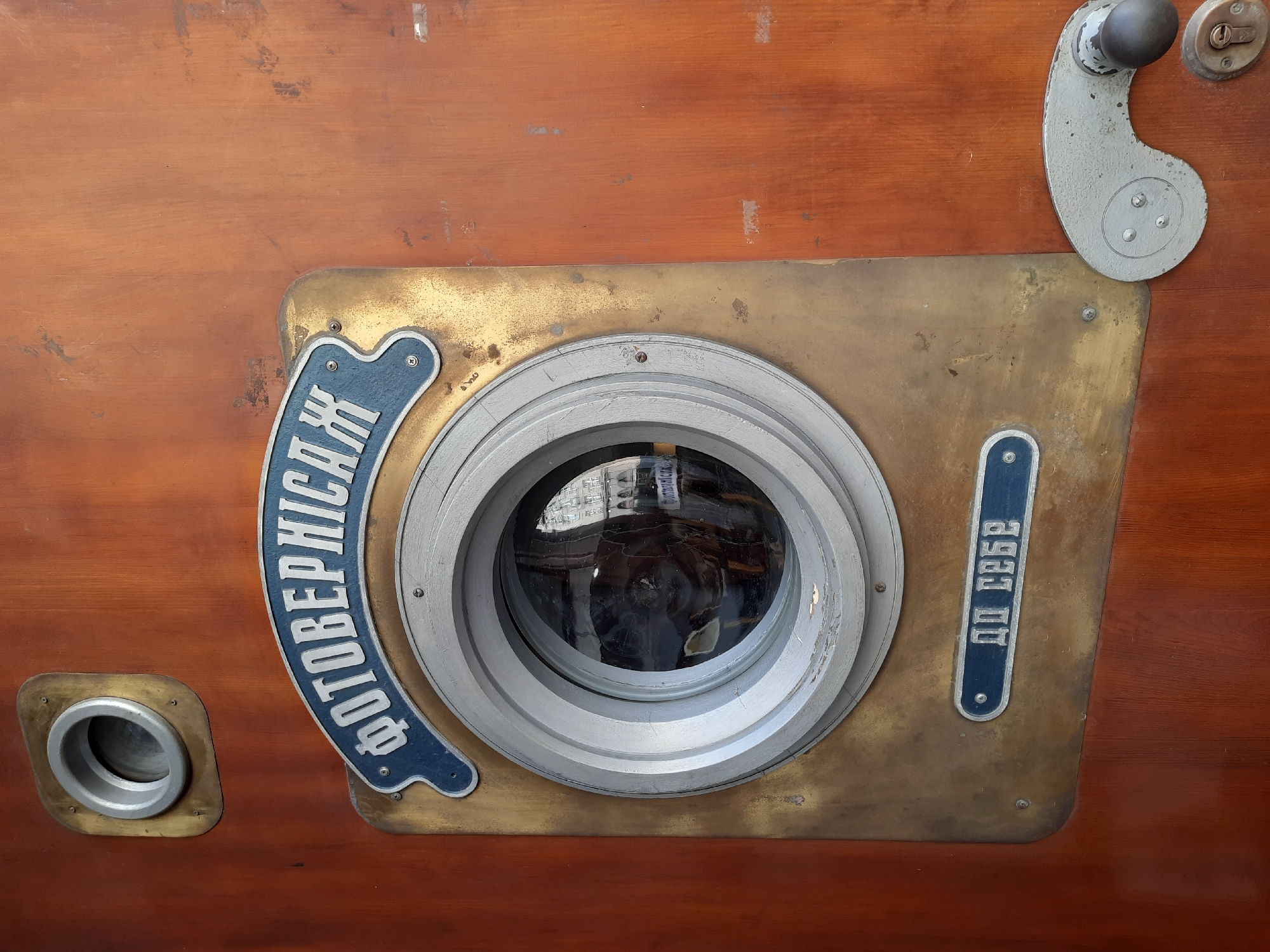 This image shows a close-up of a vintage photographic booth, likely a photobooth or a similar automated portrait service. The primary subject is the brass-plated mechanism surrounding a circular glass lens, which is the viewing window for the booth. The lens is slightly dirty, and a reflection of the interior of the booth, including what appears to be a person or a dark figure, is visible on its surface.
To the left of the lens is a smaller circular opening with a metallic rim, possibly an input slot or another control. On the left side of the brass plate, curved text in blue and white reads "ФОТОГРАФІЇ" which translates to "PHOTOGRAPHS" in Ukrainian. On the right side of the brass plate, similar blue and white text reads "до себе" which translates to "for yourself" or "yourself" in Ukrainian. These inscriptions suggest the purpose of the booth is for taking self-portraits or photographs of individuals.
The booth itself appears to be constructed of dark, polished wood. To the upper right of the main mechanism, there is a metal handle and a keyhole, hinting at the operational and security features of the device. The overall impression is one of a historical, mechanical object, likely an artifact from a past era of automated photography. The setting appears to be indoors, given the reflection in the lens and the presence of the operational controls. The lighting is not clearly discernible but seems to be ambient. The image is shot from a slightly elevated angle, focusing on the details of the photographic apparatus.