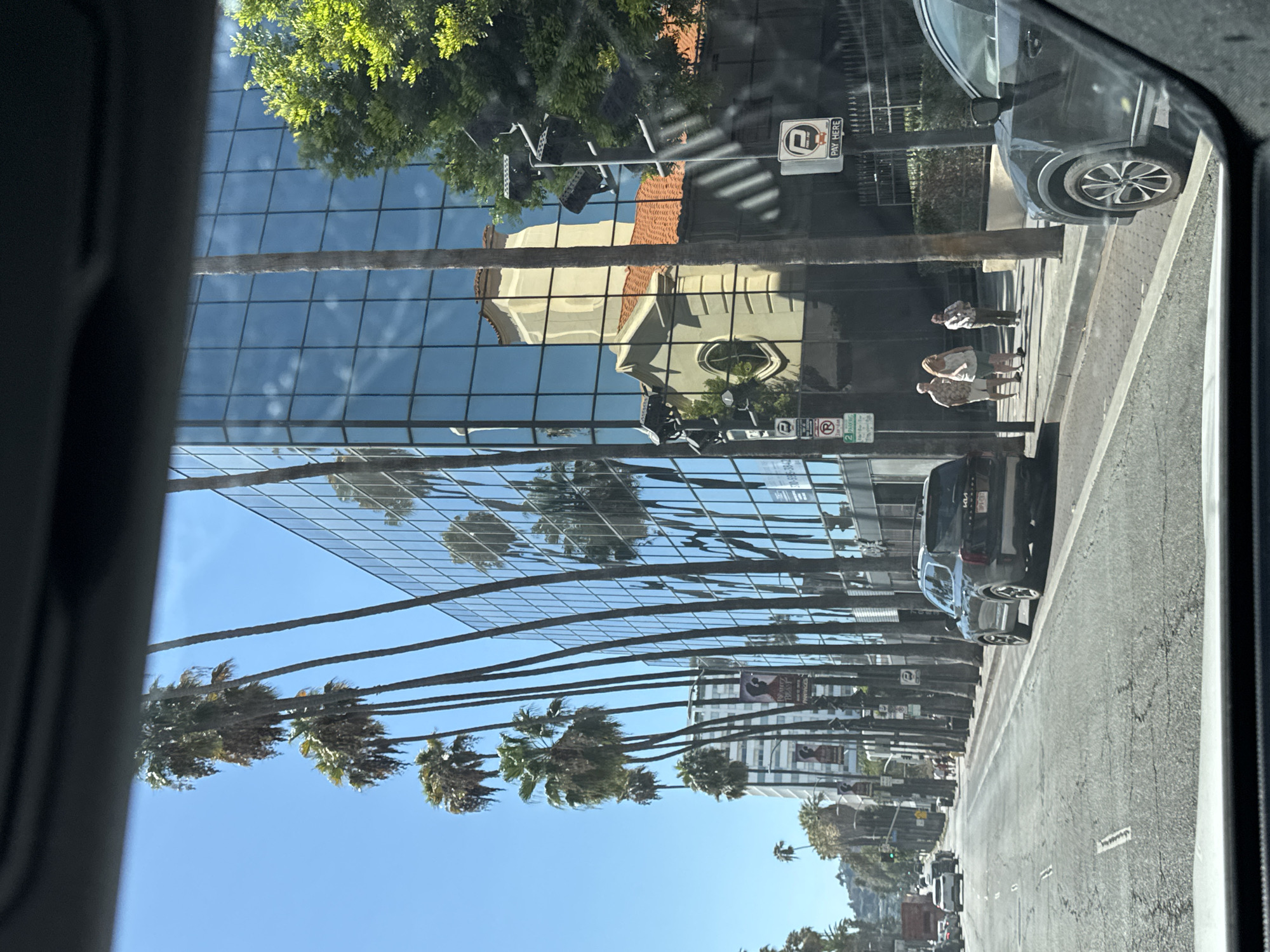 The image depicts a street scene in Los Angeles, California, USA, likely on a sunny day. A modern, glass-fronted building dominates the right side of the frame, its reflective surface mirroring the blue sky, palm trees, and a building with a terracotta-tiled roof and a bell tower. A row of tall palm trees lines the sidewalk in front of the glass building, interspersed with streetlights and signs.
In the foreground, a street is visible with several cars. A dark-colored SUV is parked on the right side of the street, and a portion of another dark-colored SUV is in the lower right corner, suggesting the image was taken from inside a vehicle. On the sidewalk, three people are walking away from the viewer.
Further down the street, traffic is visible, and more buildings and palm trees can be seen. Banners hang from poles along the sidewalk, some featuring images of women. Traffic lights and parking signs are also present. The overall impression is a typical urban streetscape in a sunny, palm-lined city.