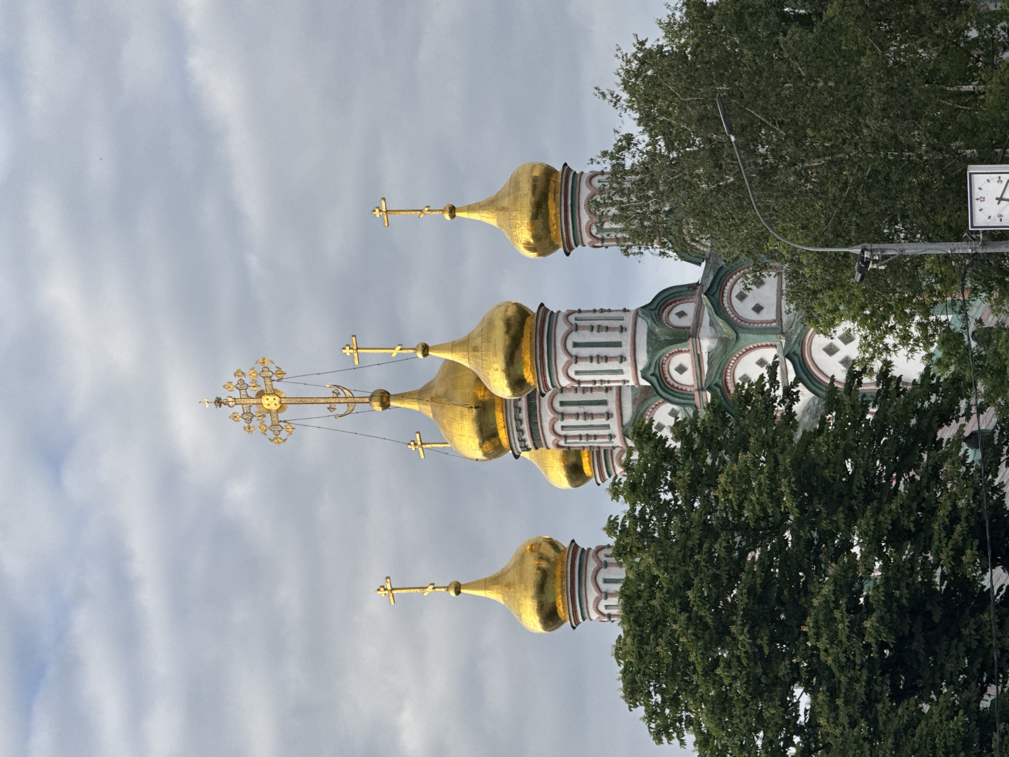 The image displays an Eastern Orthodox church, oriented as if viewed from below and slightly to the side, with the overall structure rotated 90 degrees clockwise in the provided file. When viewed upright, the church features a cluster of shimmering golden onion domes (cupolas), each topped with an ornate Orthodox cross. The most prominent cross, visible on the tallest spire to the left, is intricately designed with decorative elements.
The church's facade beneath the domes is richly adorned with decorative patterns in shades of green, white, and a reddish-brown, indicative of traditional Russian architecture. Lush green trees partially obscure the lower and right portions of the building, suggesting an outdoor, possibly park-like, setting.
To the right, a utility pole extends upwards, supporting a street lamp and a square-faced clock. The clock face, showing numbers 12, 3, 6, and 9, clearly indicates the time as approximately 10:45. The sky is overcast with soft, light gray clouds, suggesting a cloudy but well-lit day. No people are visible, and thus no specific activity or emotions are depicted. The architectural style, particularly the onion domes and Orthodox crosses, strongly cues a location in Russia, specifically Moscow as per the provided context.