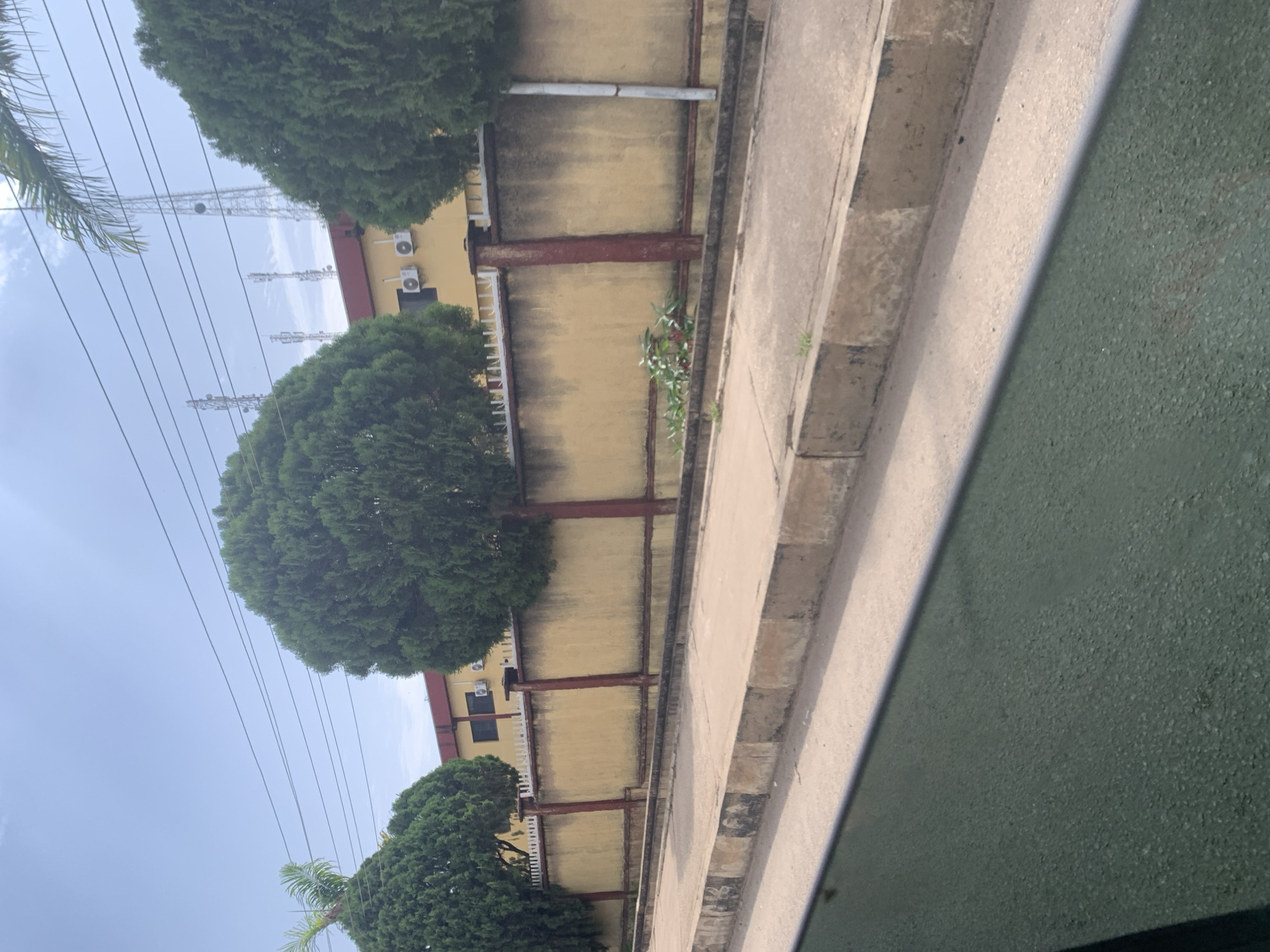 The image depicts a street scene in Jos, Nigeria, characterized by a yellow wall topped with a white spiked fence, separating a paved area from a building. Several cypress trees with rounded canopies are positioned along the wall. The building behind the wall is yellow with a red roof and has air conditioning units mounted on its exterior. In the background, communication towers are visible against a sky with scattered clouds. A concrete sidewalk runs in front of the wall, and a street with a green tinted foreground, possibly from a vehicle's window, is also present. The overall impression is one of a moderately developed urban or suburban environment.