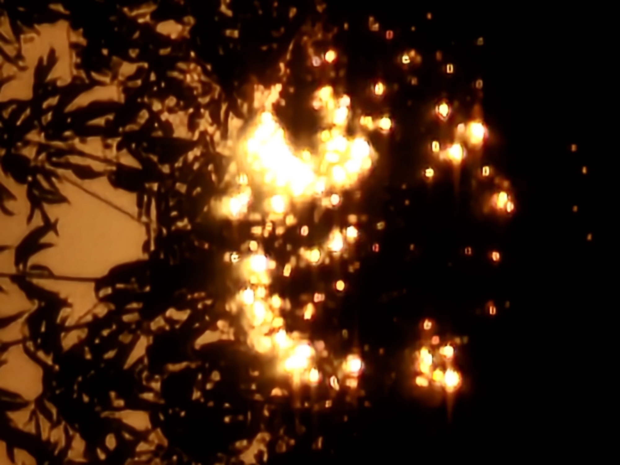 The image shows a dark scene with blurry lights. In the upper portion, silhouetted foliage is visible against a warm, yellowish background. Below this, numerous bright, out-of-focus points of light are scattered throughout the frame, appearing as orbs and flares of yellow and orange. The foreground and lower sections are predominantly black. There is no visible indication of the city of Khimki, Russia, or any specific urban environment. The scene appears to be an abstract or artistic representation, possibly of lights at night, bokeh effects, or a visual representation of heat or energy.