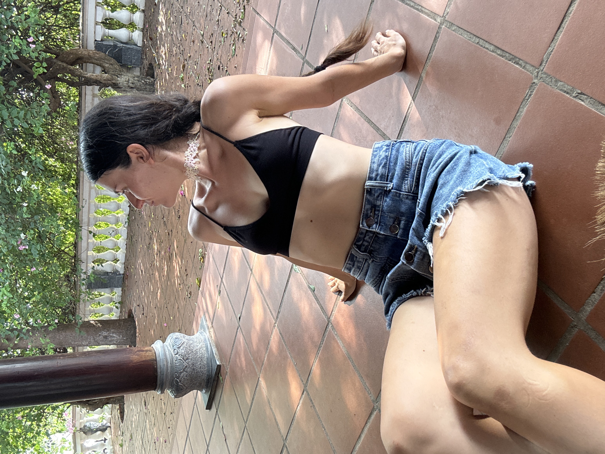 In Mũi Né, Vietnam, a woman with dark hair in a braid sits on a tiled patio, her legs extended. She wears a black bikini top and denim shorts with frayed edges. Her left arm rests on the ground behind her, while her right arm is bent at the elbow, with her hand near her knee. A white floral choker adorns her neck. The patio floor is made of reddish-brown square tiles. Behind her, a large, dark wooden pillar stands to the left. In the background, a white balustrade lines the edge of the patio, with trees and lush green foliage beyond. Sunlight filters through the leaves, casting dappled shadows on the tiles.