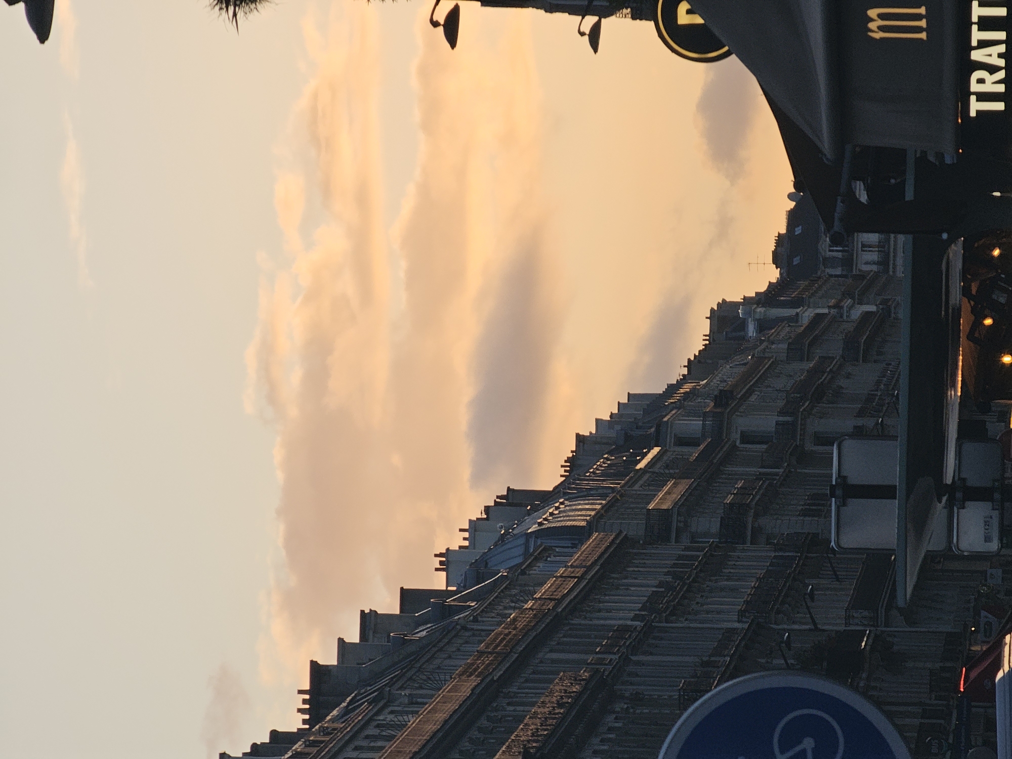 The image depicts a cityscape in Paris, France, captured during the sunset. In the foreground, a restaurant or café's sign reading "TRATTORIA" is visible. The buildings, with their characteristic Parisian architecture, stretch into the distance, their roofs forming a repetitive pattern against the backdrop of the sunset-lit sky. The clouds are tinged with shades of orange and pink, indicating the sunset. Street signs and possibly traffic lights are interspersed within the urban scene. The image is slightly rotated clockwise.
