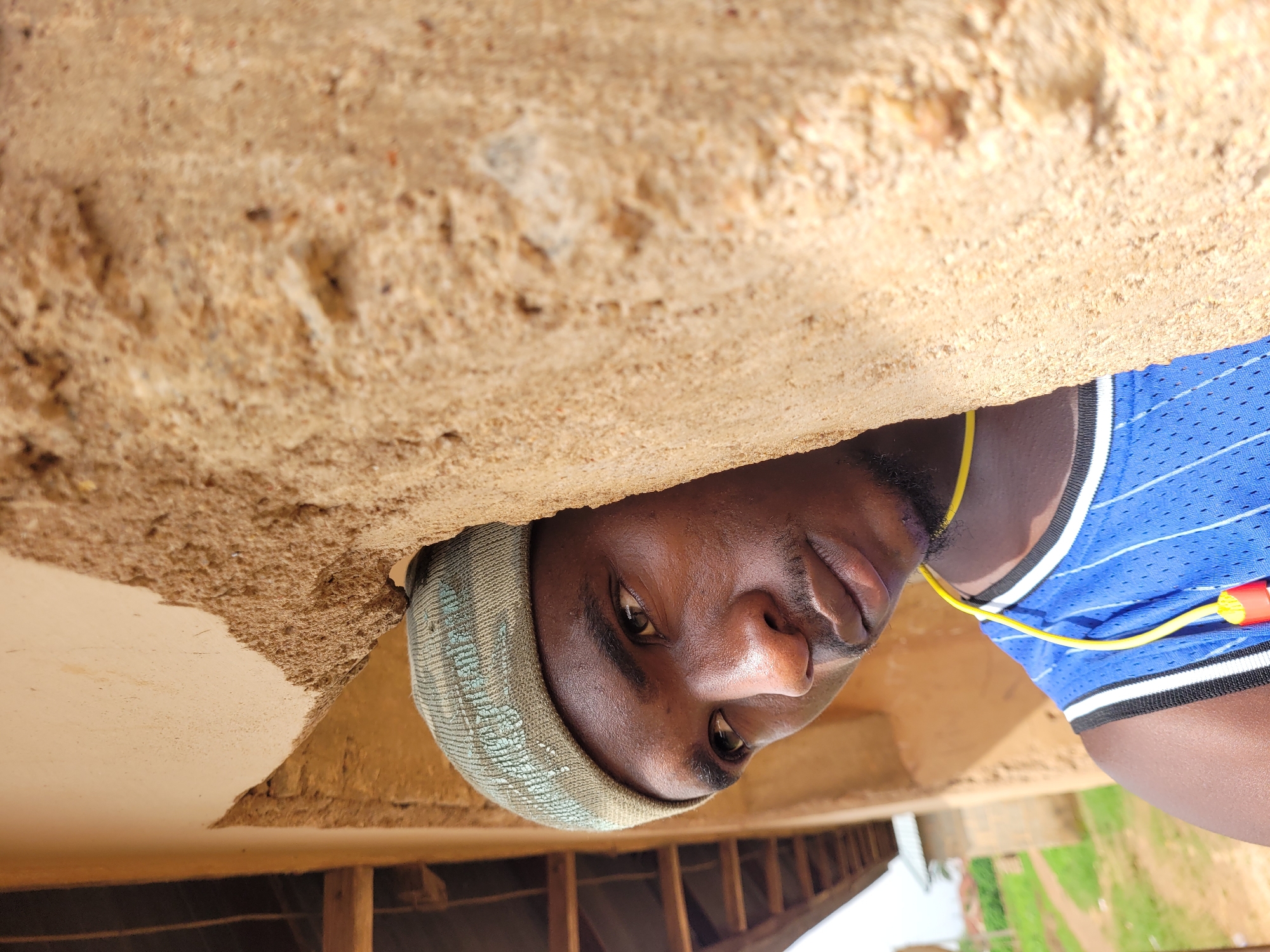 The image shows a close-up, outdoor, waist-up portrait of a young Black man in Bukuru, Nigeria. He is positioned to the left, looking to the right, partially obscured by a rough, tan-colored concrete pillar. He is wearing a light green knit cap with a subtle patterned texture, and a blue athletic tank top with white pinstripes and black and white trim. A bright yellow cord, likely from earphones, is visible around his neck, with red and yellow earpieces hanging down in front of his chest. The background, visible to the left and behind him, shows a wooden roof structure and hints of green vegetation, suggesting a rural or semi-urban setting. The lighting indicates it is daytime, with sunlight illuminating the scene.