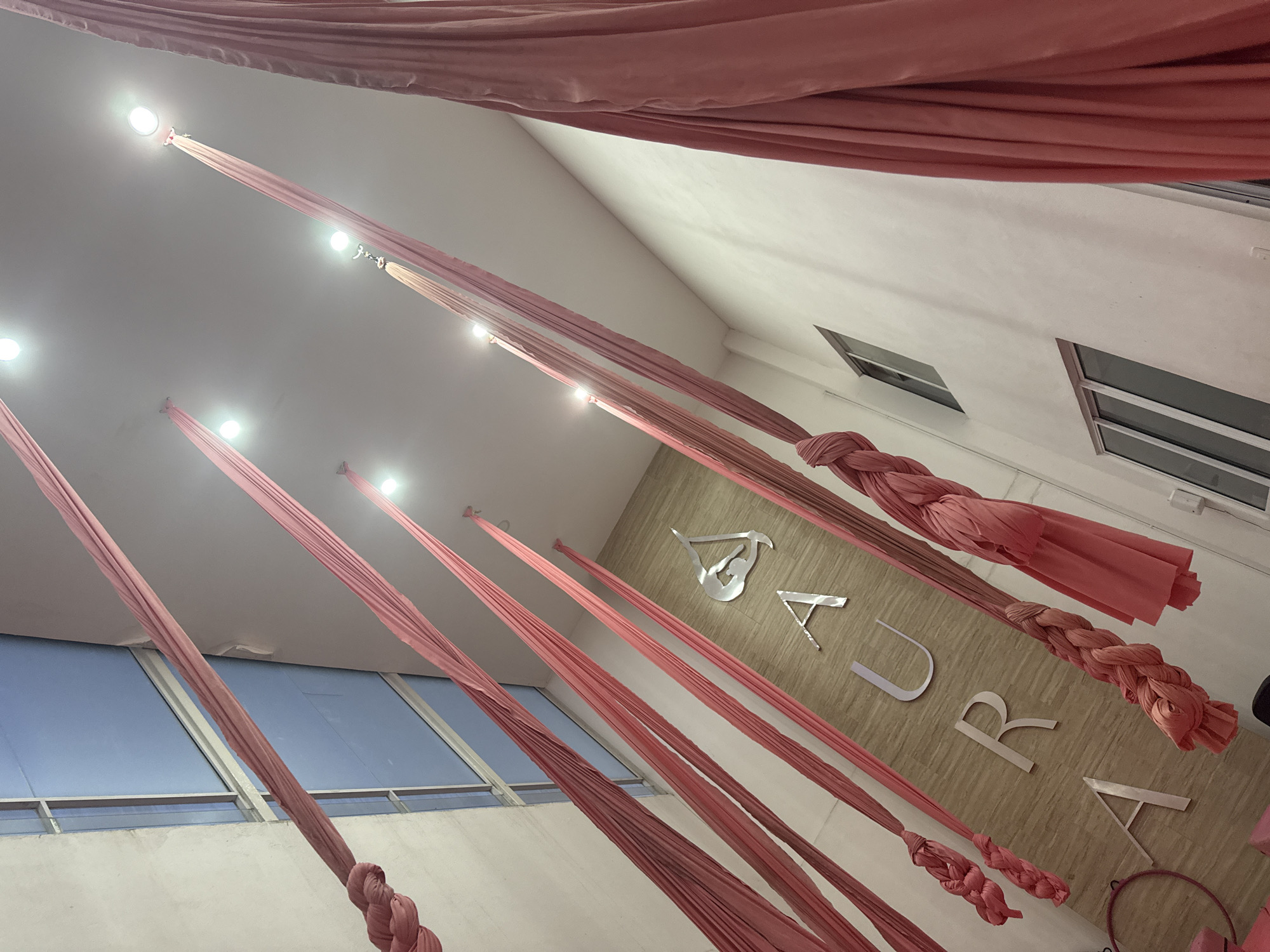 The image shows the interior of a studio, possibly in San Andrés Cholula, Mexico, judging by the decor. Several lengths of rose-colored fabric hang vertically from the ceiling, where small lights are attached at their points of suspension. Some of the fabric lengths are knotted at the bottom. The word "AURA" is displayed prominently on the wall, with silver letters. There are windows visible in the background.
