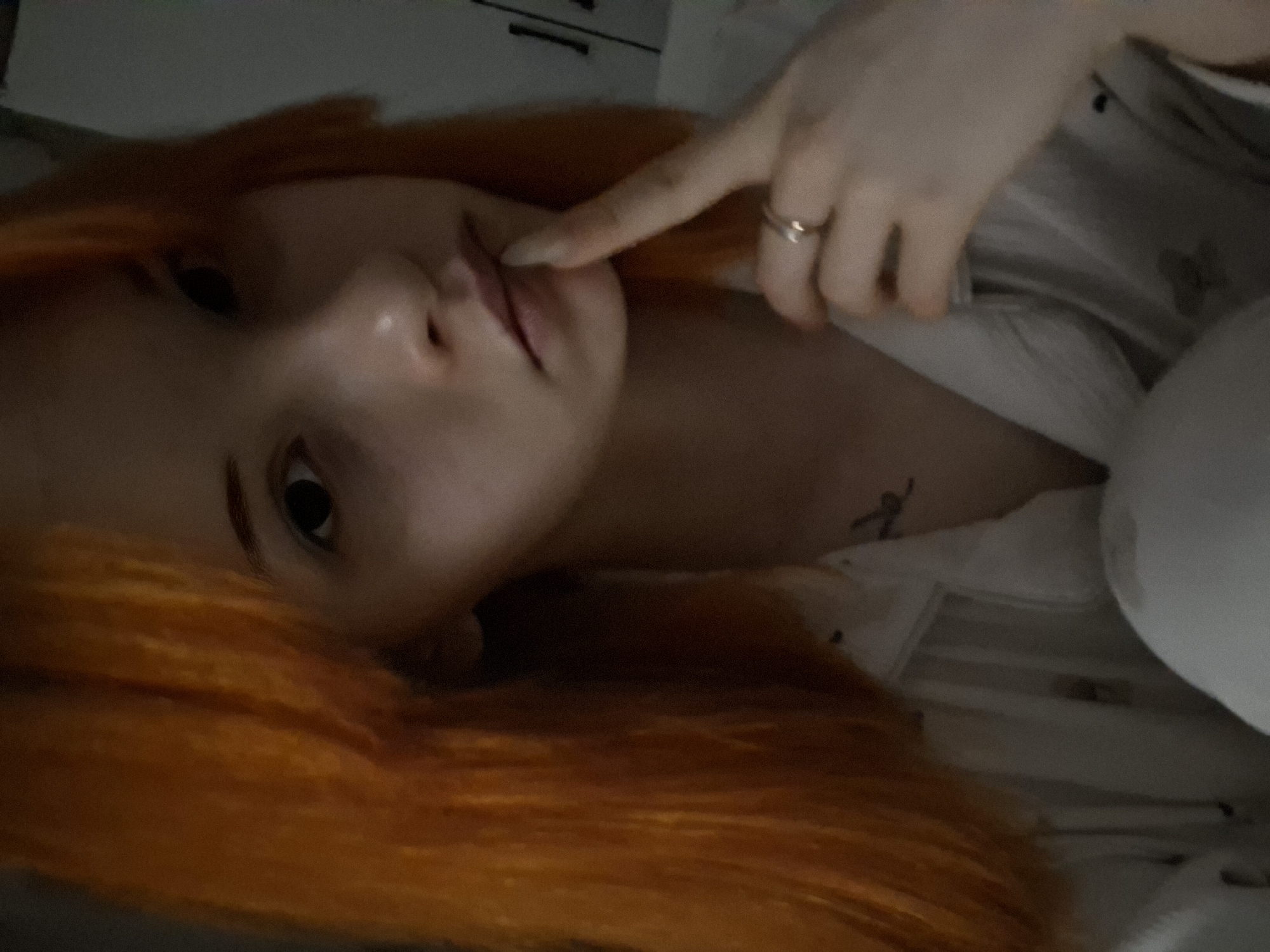 A close-up, low-light photograph captures a person with distinctive bright orange hair lying down, looking towards the camera. The subject's face is partially obscured by their hand, with one finger gently touching their lips. A simple ring adorns the finger.
The person's dark eye and part of their nose are visible, along with a fair complexion. Their long, vibrant orange hair spreads around their head, suggesting a relaxed posture. A small, dark tattoo, appearing to be a minimalist figure or symbol, is visible on their neck or upper shoulder area.
They are wearing light-colored clothing, possibly striped pajamas or a casual top, which is partially visible around their shoulder and neck. The background is dark and indistinct, with faint shapes that could be room furniture, indicating an indoor setting, likely a bedroom. The dim lighting suggests it could be evening or nighttime. The overall mood is quiet and intimate. There is no visible text in the image.