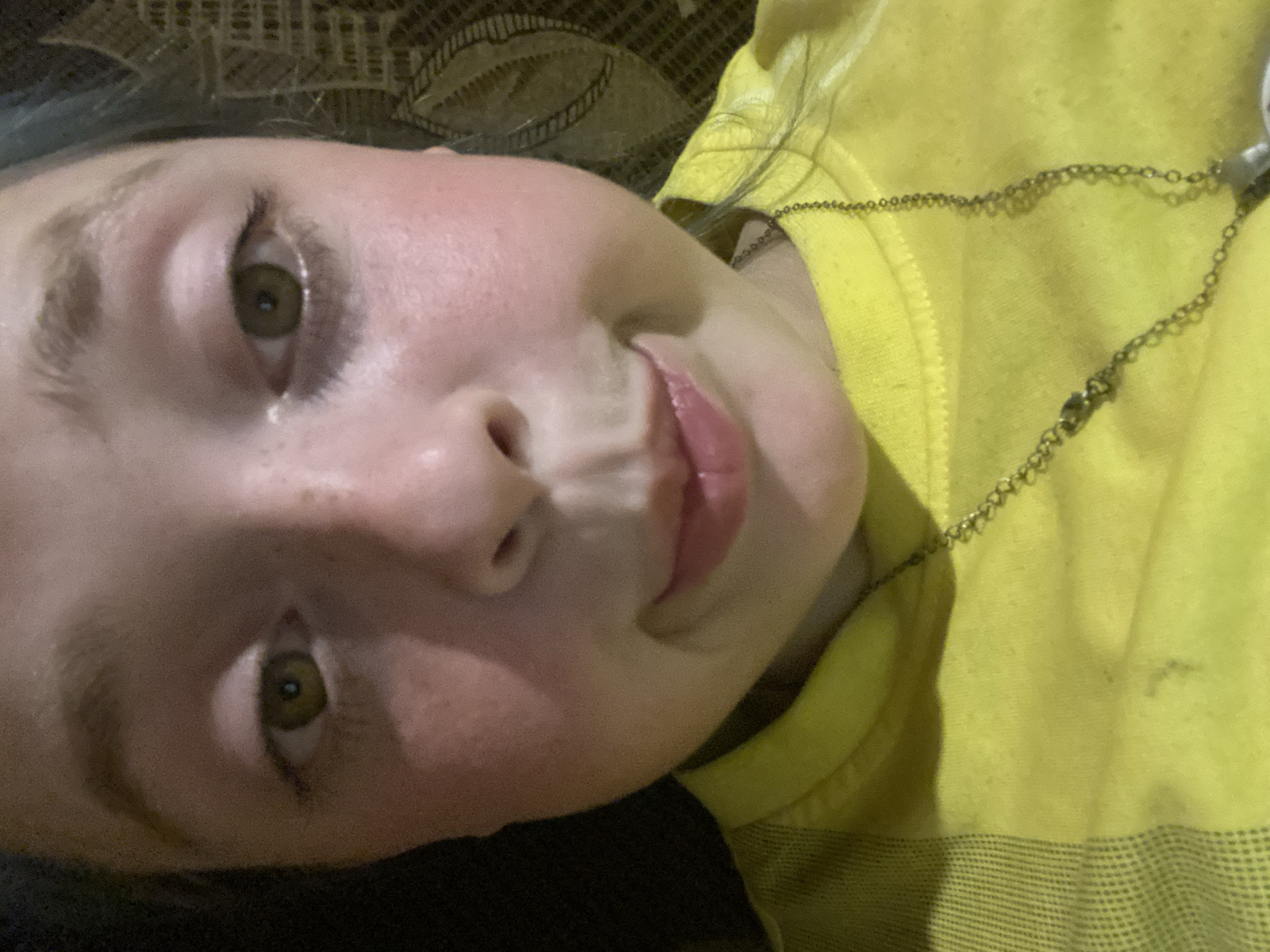 The image is a close-up, selfie-style photograph of a young person with light skin and freckles. They have large, greenish-brown eyes and a slight pout on their lips. The person is wearing a bright yellow t-shirt and a delicate silver chain necklace. The background is dark and out of focus, with some texture suggesting a woven material or patterned upholstery. The lighting is somewhat dim, casting shadows on the face and highlighting the contours of the cheeks and nose. The scene appears to be indoors. There is no information in the image to indicate the location in Nova Praha, Ukraine.