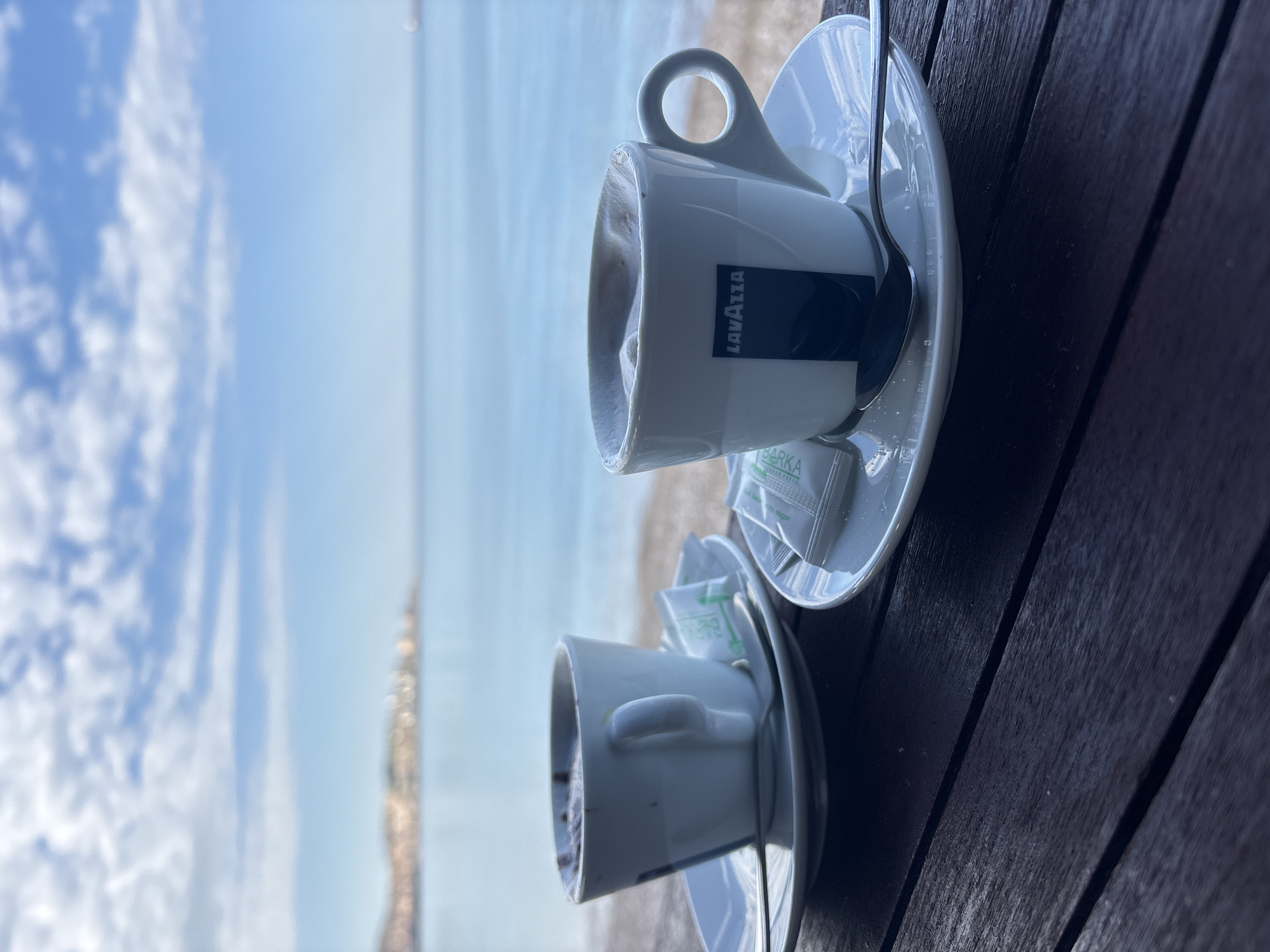 Two cups of Lavazza coffee sit on a dark wooden table. The cups rest on white saucers and each has a small spoon and a sugar packet. In the background, a blurred scene shows the sea and sky, suggesting a beachside location. Based on the visual context, this is most likely in Bar, Montenegro.