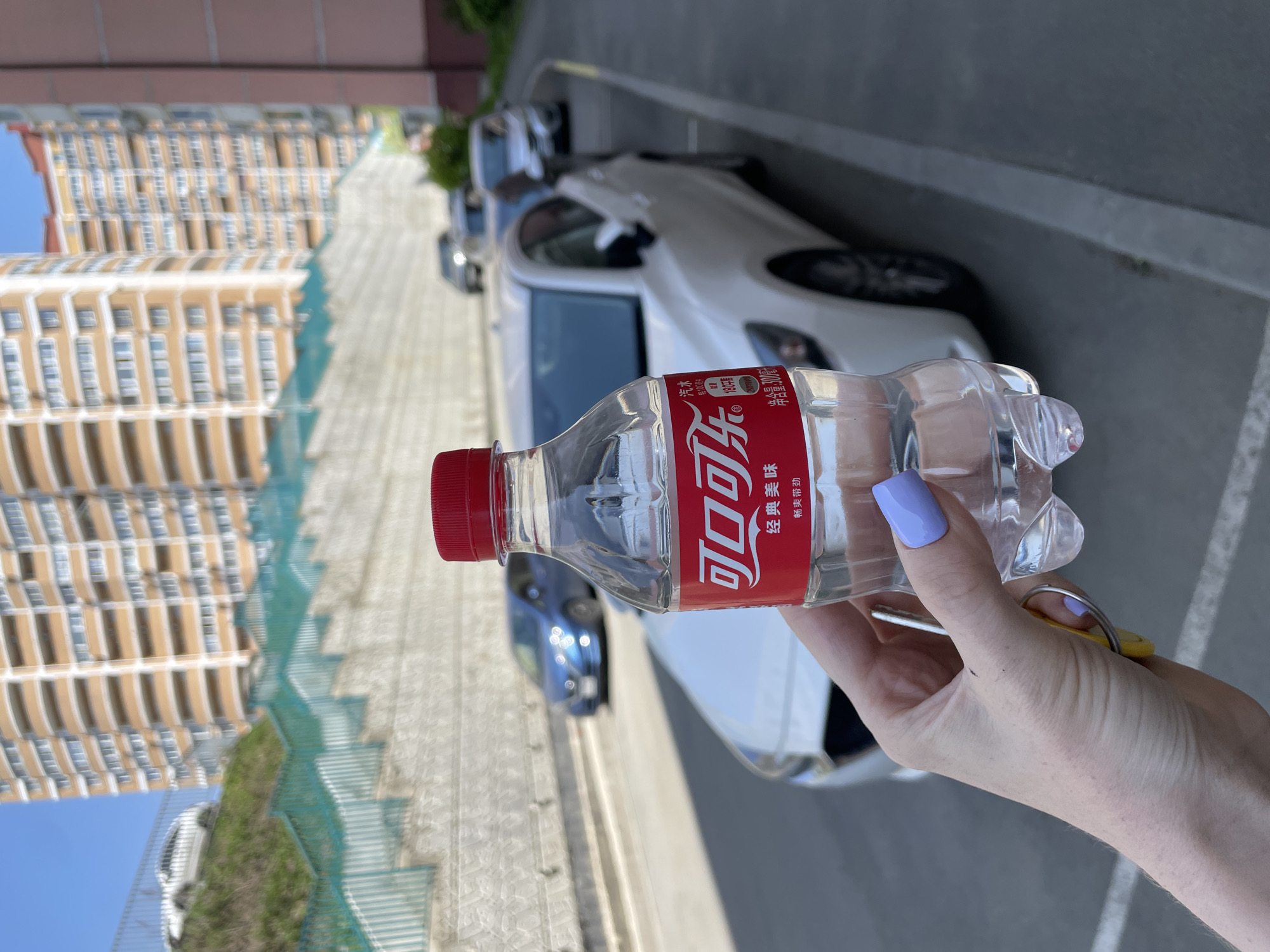 The image, taken in bright daylight, features a close-up of a person's right hand holding a small plastic bottle and a keychain. The hand, with light purple nail polish on the index finger, grips a clear bottle with a red screw-cap and a striking red label. The label prominently displays white Chinese characters: "可口可乐" (Coca-Cola), alongside "经典美味" (Classic Delicious) and "净含量:300ml" (Net Content: 300ml). The bottle contains a clear liquid, likely water. A yellow keychain with a silver key is also held by the same hand.
Behind the hand and bottle, a paved parking area is visible, with several cars parked, including a white car positioned directly behind the bottle. Further back, a tall, multi-story building with a light brown or beige facade and numerous windows stands under a clear blue sky, suggesting sunny weather. A teal-colored, mesh-like fence runs along the edge of the paved area, interspersed with some green foliage. The scene depicts an urban setting, consistent with Vladivostok, Russia, as provided in the context, and the Chinese text on the bottle hints at regional commercial influences.