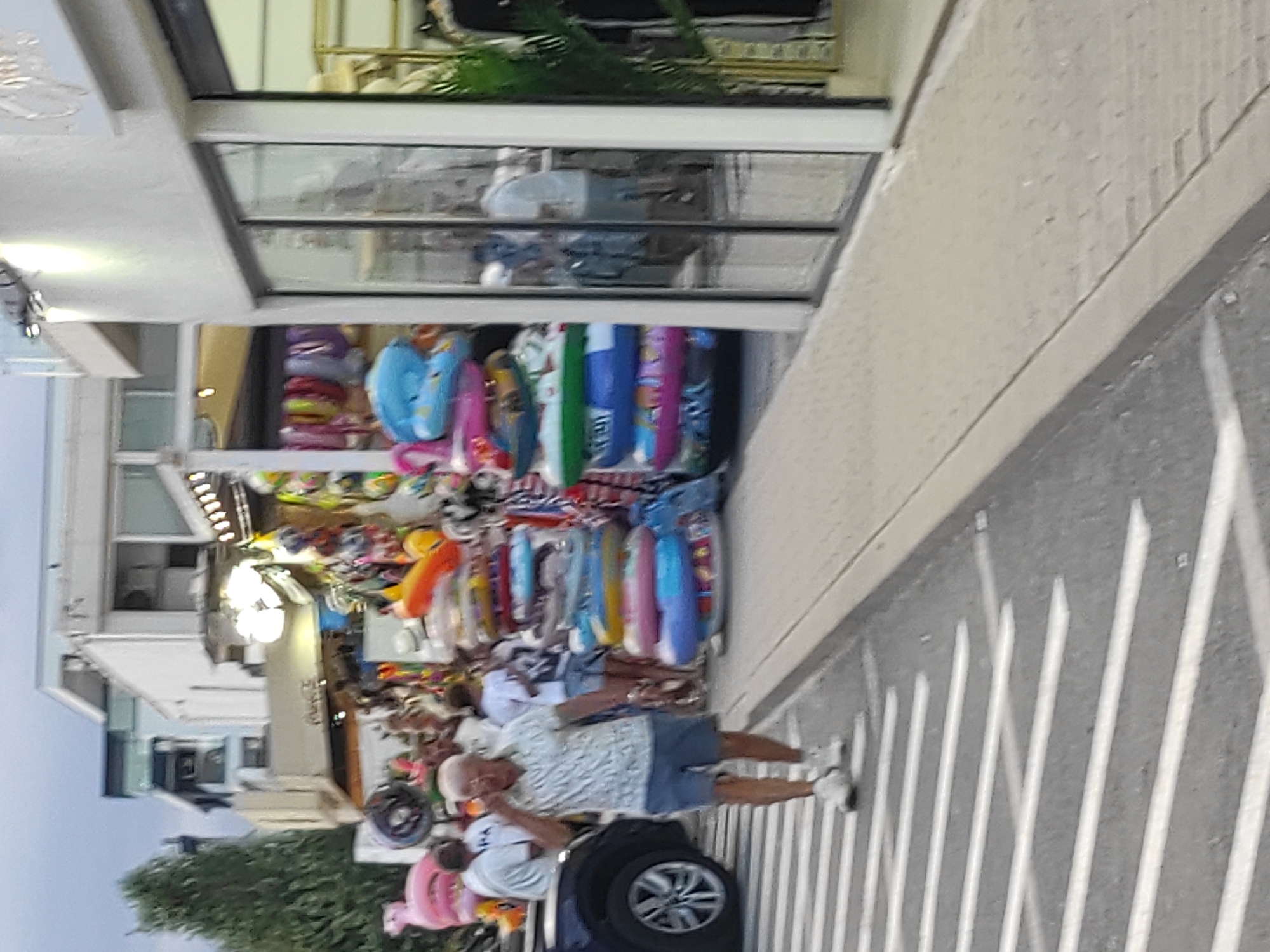 The image depicts a street-level view of a shop displaying a variety of inflatable beach toys, such as swim rings and pool floats, arranged in rows. The shop appears to be an outdoor stall or an open-front store, with merchandise spilling out onto the sidewalk. In the foreground, two people are visible. One, an elderly man wearing a patterned short-sleeved shirt and blue shorts, is standing and looking towards the viewer, with his hand near his face as if touching it. He is wearing white sneakers. Behind him and to his left, another person, wearing a light-colored short-sleeved shirt and denim shorts, is also visible, appearing to be standing. The overall scene suggests a warm, possibly summer day, with the abundance of beach items. The setting seems to be a tourist or coastal area, given the merchandise. The image is taken from a slightly angled perspective, and the lighting suggests it could be late afternoon or early evening, with a bright light source from the shop's interior illuminating some of the displays. There is no visible text in the image. The location is Năvodari, Romania, a town known for its proximity to the Black Sea coast.