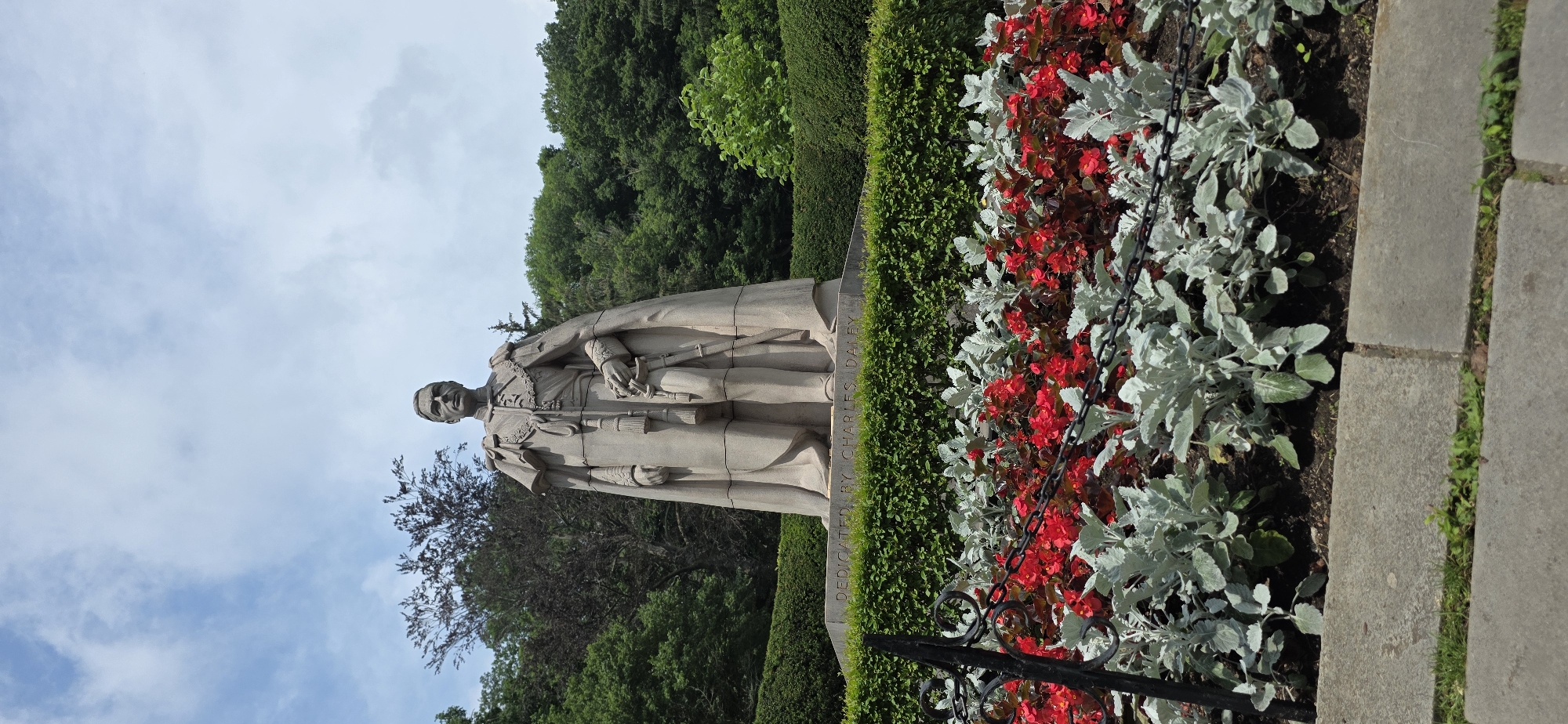 A stone statue of a man in regal attire stands in front of a dense hedge and lush green trees under a partly cloudy sky. The statue depicts a man wearing a long cape over a military uniform, complete with medals and a sword at his side. His hands are clasped in front of him. The base of the statue is inscribed with the words "DEDICATED BY CHARLES DALEY". In the foreground, a bed of bright red flowers is interspersed with silvery-green foliage, separated from the statue by a low, manicured hedge and a black metal fence with a chain. The statue is located in Niagara Falls, Canada, as evidenced by the inscription on the base.