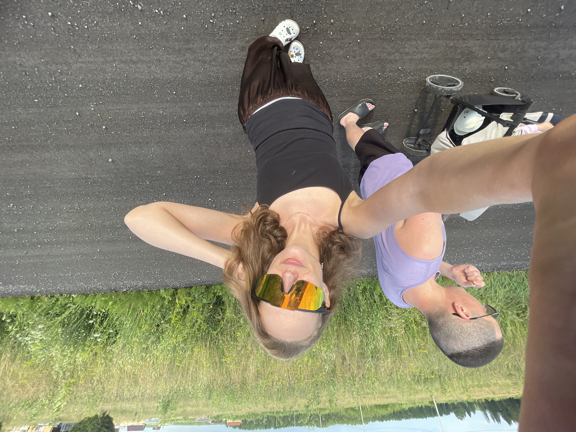 In Подшивалово, Russia, a man and a woman are taking a selfie outdoors. The man, with a shaved head and sunglasses, wears a purple tank top and dark shorts. He appears to be pushing a stroller. The woman, in the foreground, wears a black tank top, brown pants, and white Crocs with charms. She has on large, reflective sunglasses and is touching her hair. The ground is a dark, gravelly road, and the background is a field of tall, green grass with some houses and trees visible in the distance under a partly cloudy sky.