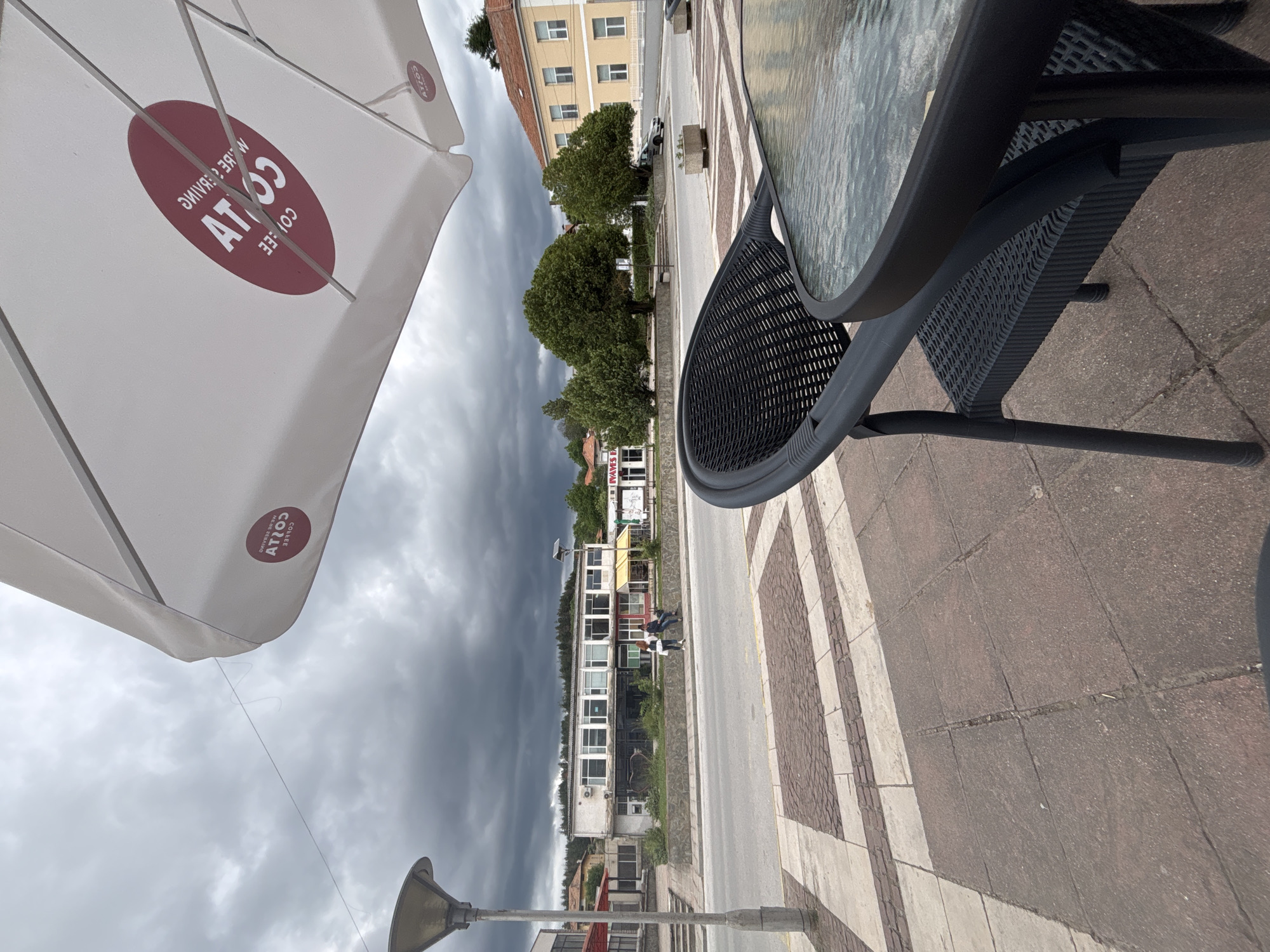 The image depicts a view from an outdoor cafe seating area in Garmen, Bulgaria, under a heavily clouded sky. In the foreground, a black rattan chair and a glass-topped table are visible. An open white umbrella with a red "Costa Coffee" logo is tilted overhead. Beyond the cafe area, a street runs horizontally across the frame, with a sidewalk and pavement in front of it. Across the street, several buildings are present, including a long, light-colored building with multiple windows, a yellow building with red trim, and other structures further in the distance. A few trees are scattered throughout the scene. Two individuals are seated on a low wall in front of one of the buildings. The overall atmosphere is overcast and slightly subdued.