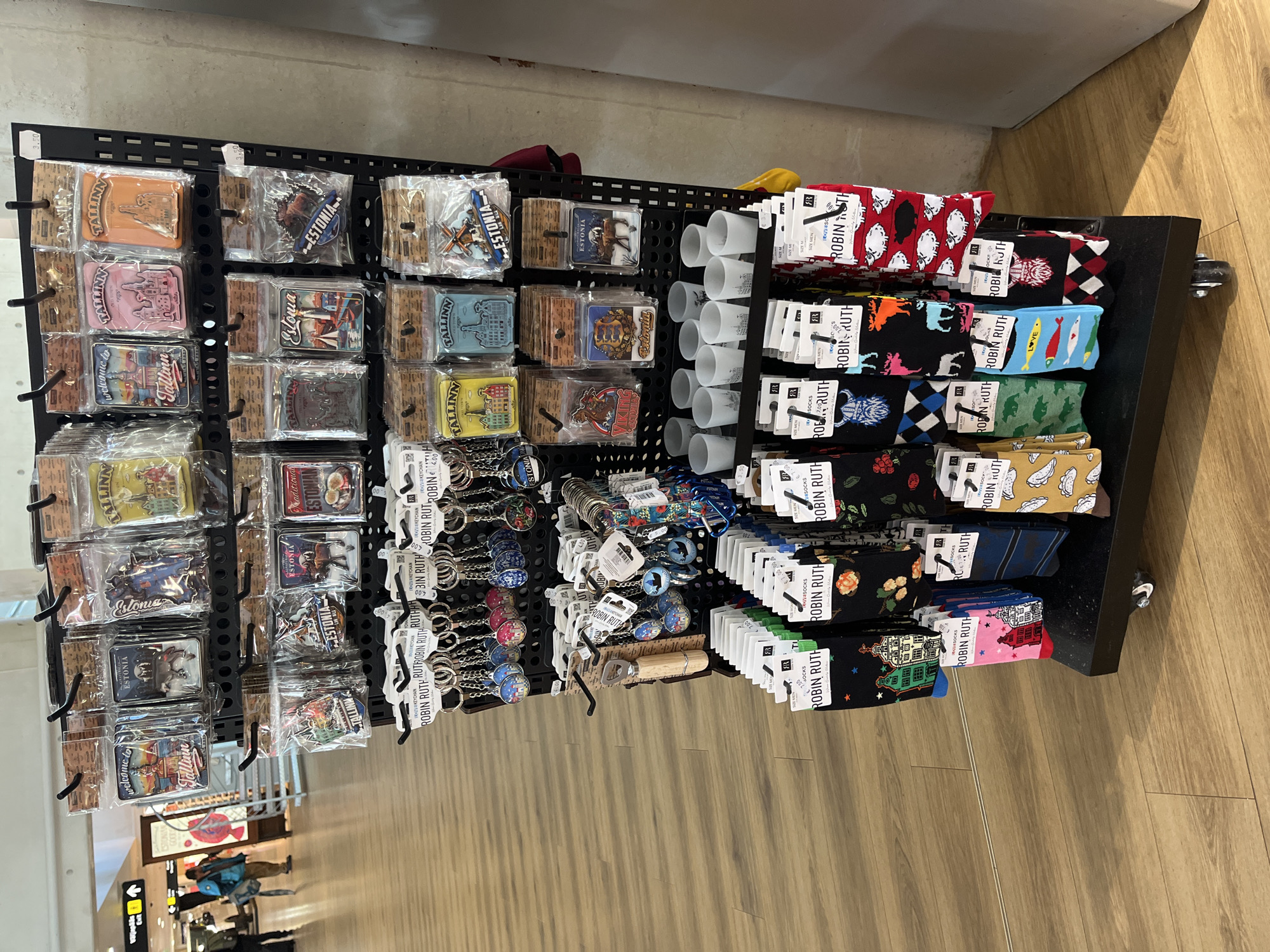 A retail display rack is positioned on a light wood floor in what appears to be an indoor commercial space, possibly an airport or duty-free shop. The rack, which is black and has wheels, is filled with merchandise. The top two shelves of the rack feature souvenir magnets and keychains, predominantly with designs related to Tallinn, Estonia, including images of the city skyline, windmills, and local wildlife. There are also rows of small frosted glass shot glasses with silhouette designs of Tallinn buildings. The bottom shelves are stocked with pairs of patterned socks, also featuring designs with a Tallinn theme, such as buildings, animals, and traditional patterns. The background shows a section of a building with a textured concrete wall, and further in the distance, a commercial area with signage and people, suggesting a public transit hub or shopping center.