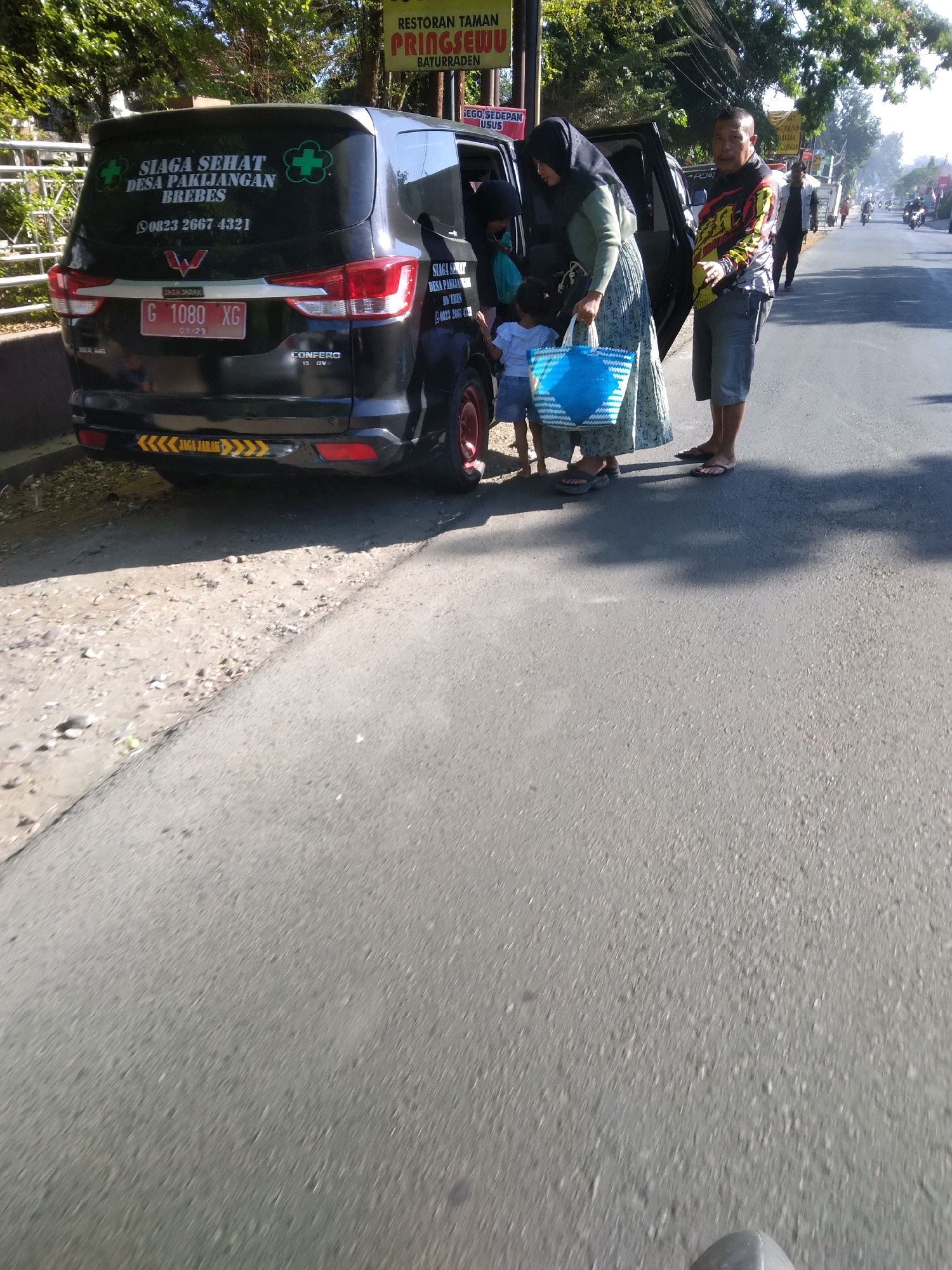 This is a medium-angle, eye-level, outdoor shot of a black van with the words "SIAGA SEHAT DESA PAKIJANGAN BREBES" on its rear window, indicating it's a health service vehicle for the village of Pakijangan in Brebes. The van is parked on the side of a paved road. Two adults, a woman in a headscarf and a long skirt, and a man wearing a colorful long-sleeved shirt and shorts, are standing next to the open rear passenger door of the van. The woman is holding a large blue and white woven bag. A young child in a blue shirt and shorts is standing near the woman, reaching into the van. The man is looking towards the camera. The road appears to be in a somewhat rural or suburban area, with trees lining the left side and other vehicles and people visible in the background further down the road. There is a sign visible on a post behind the van. The overall lighting suggests it is daytime with bright sunlight creating shadows. The scene is focused on the van and the people interacting with it.