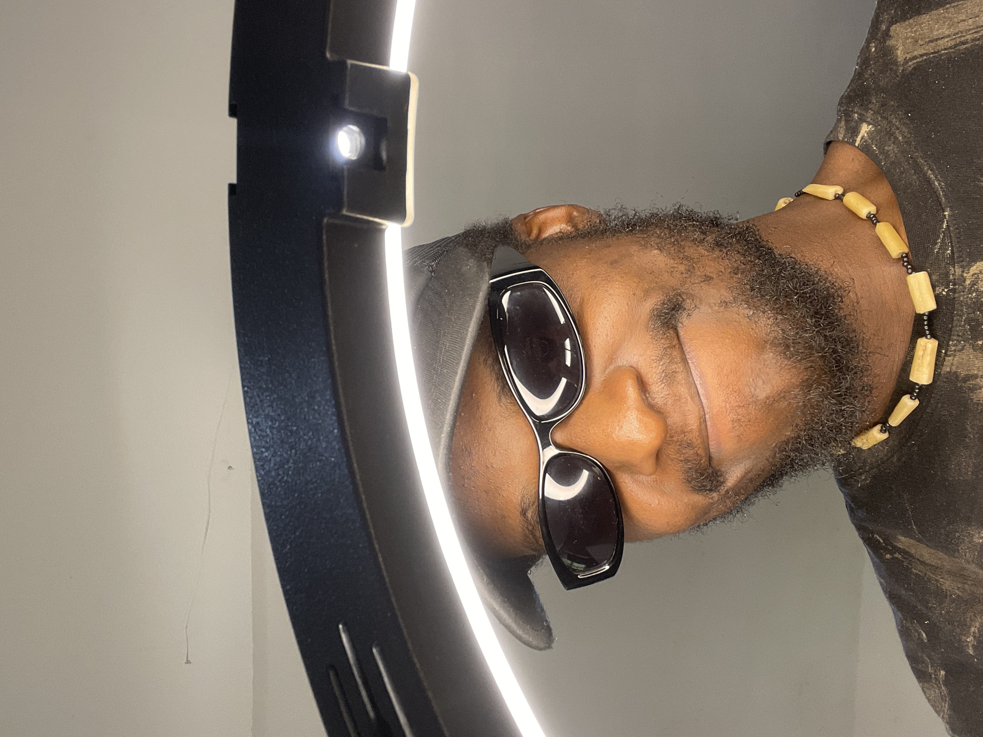 A medium shot captures a Black man wearing sunglasses and a baseball cap, posing against a plain, neutral background. He is also adorned with a necklace made of light-colored wooden beads. The lighting suggests an indoor setting, possibly a studio, with a ring light prominently positioned above and behind him, casting a soft, even illumination. The man is looking towards the camera with a subtle smile, his head tilted slightly. The overall impression is a casual and confident portrait. The location in Abuja, Nigeria, is not discernible from the visual elements of the image.