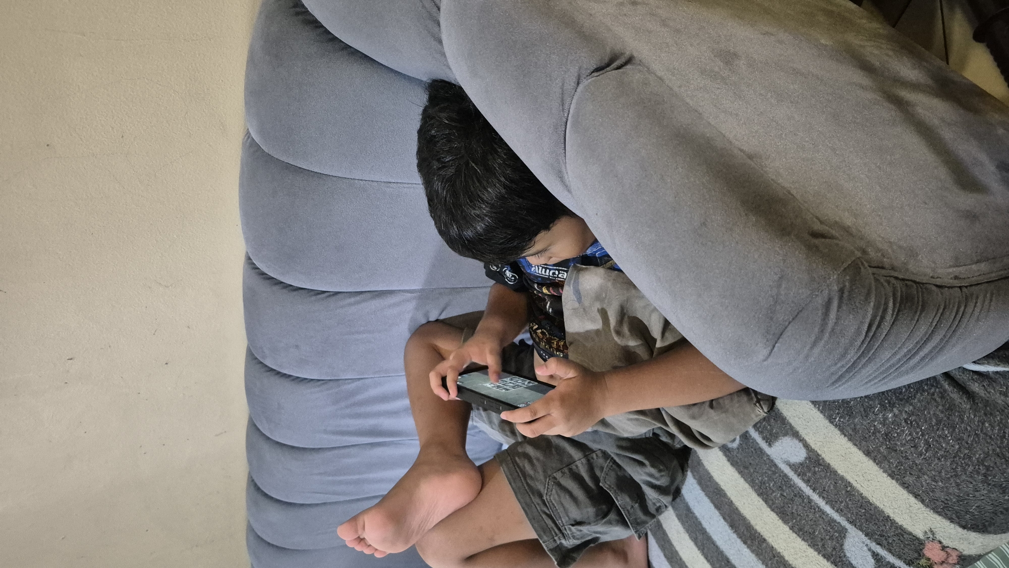 A child lies on a blue couch, playing on a mobile phone. The child is wearing a black t-shirt with a graphic design and gray shorts. The couch is in a room with a light-colored wall. The child's feet are propped up on the back of the couch. A striped blanket is visible beneath the child. The scene is likely taking place in a residential area of South Tangerang, Indonesia.