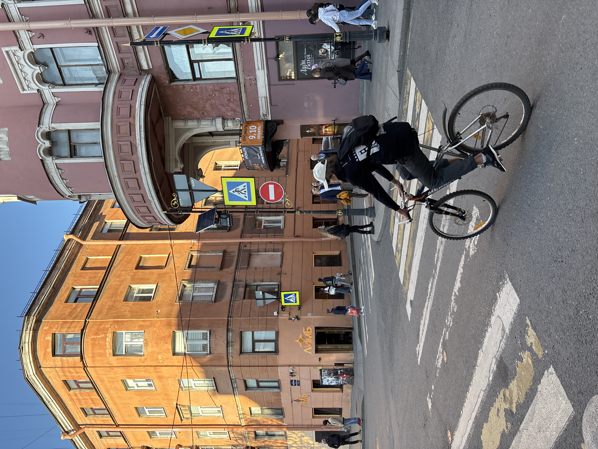 A street scene in Saint Petersburg, Russia shows a bicyclist riding across a pedestrian crosswalk. The buildings are multi-story and have a golden hue, except for one on the upper left which is pink. Pedestrian crossing signs are posted on poles near the crosswalk. People are walking along the sidewalk, and a cafe or bar called "Olymp" is visible on one of the buildings.