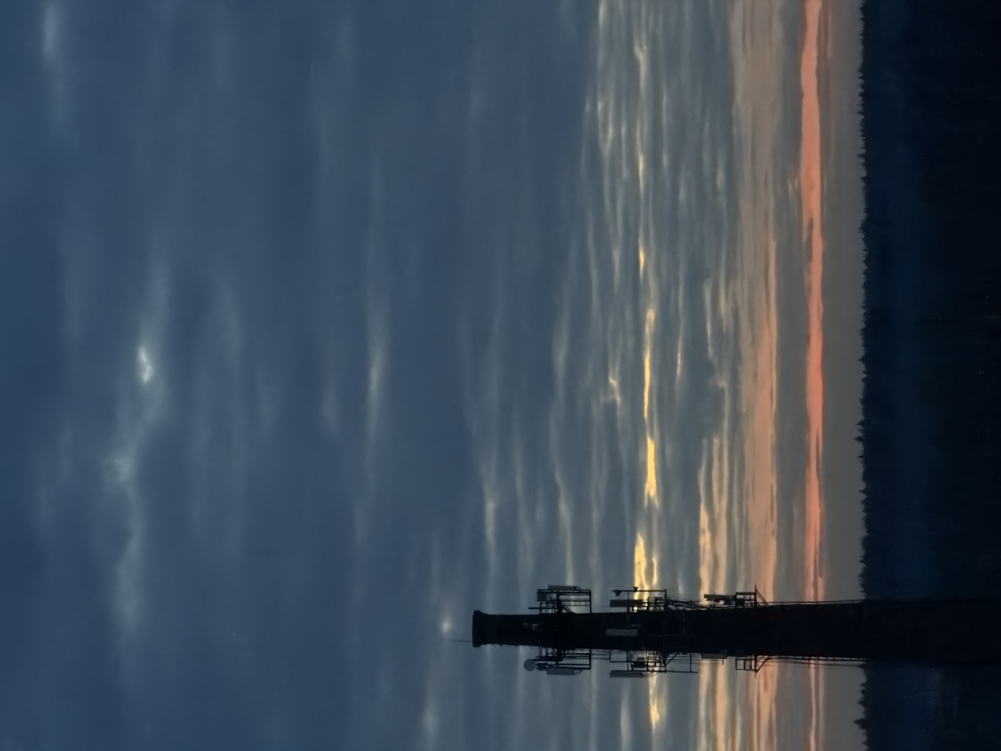 A communications tower is silhouetted against a cloudy sky, reflecting in the water below. The sky transitions from blue to orange near the horizon. A dark treeline is visible on the right edge of the image. Location: Unknown, Unknown.