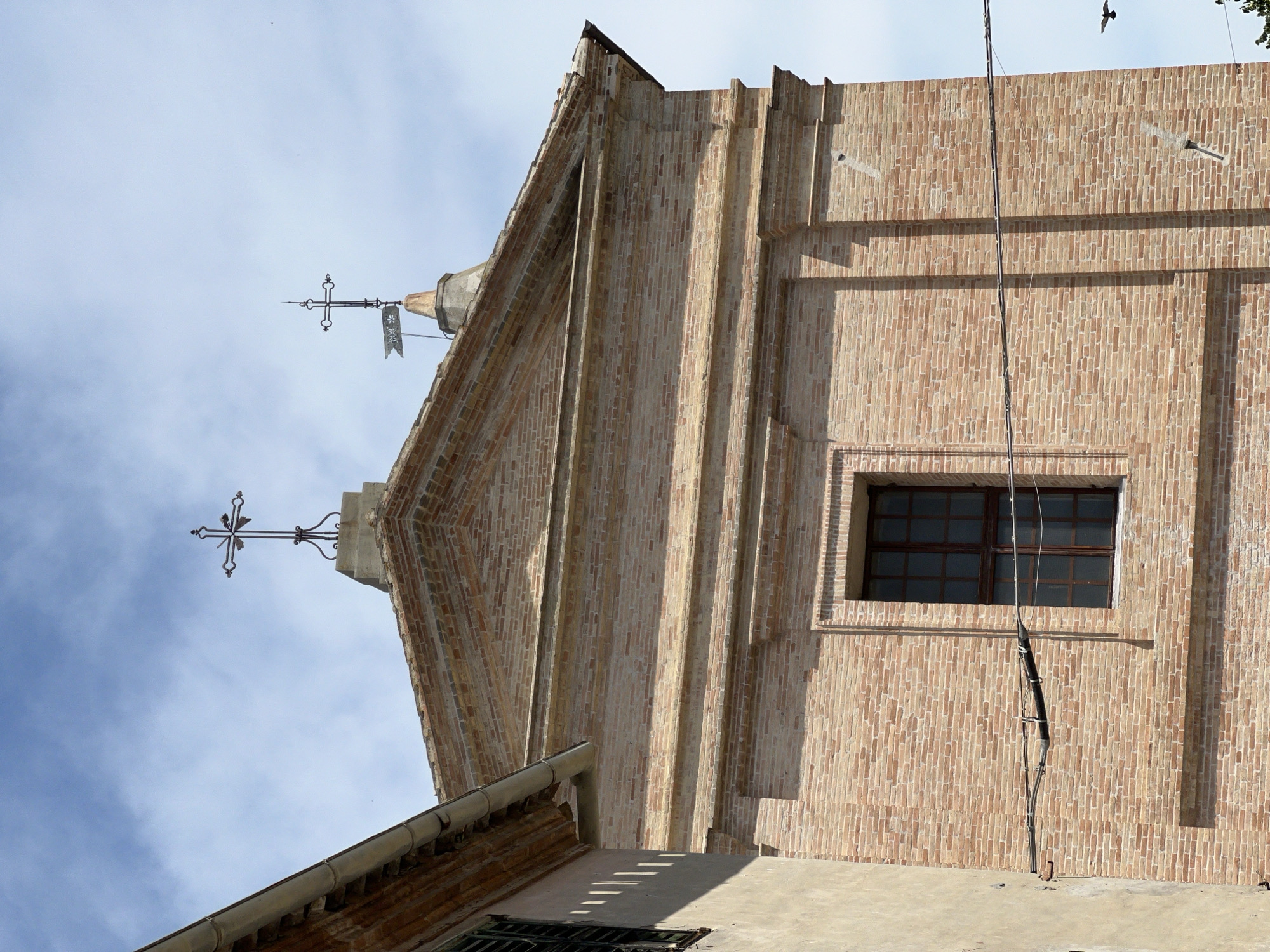 The image shows the facade of a brick building, likely a church, in Barbara, Italy. The building has a pitched roof with two crosses on top, one of which is topped with a flag. There is a single window on the facade, with a dark frame and multiple panes. The building is made of reddish-brown bricks laid in a stretcher bond pattern. A smaller, lighter colored building is partially visible on the left, with a gutter running along its edge. The sky is cloudy and blue. A bird is visible in flight in the lower right quadrant of the image. Electrical wires run horizontally across the facade.