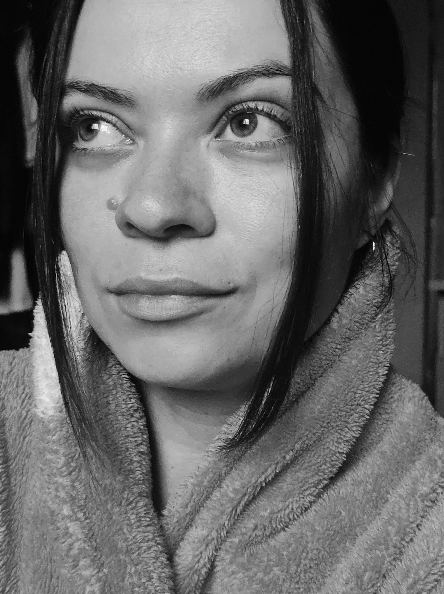 A black and white portrait depicts a woman with dark hair and fair skin. Her hair is styled with loose strands framing her face, pulled back from her forehead. She has a mole on her upper nose area. She appears to be wearing a soft, collared robe. Her gaze is directed upwards and to the left. A small hoop earring is visible in her left ear. The background consists of blurred dark shapes, possibly furniture. The location and city are unknown.