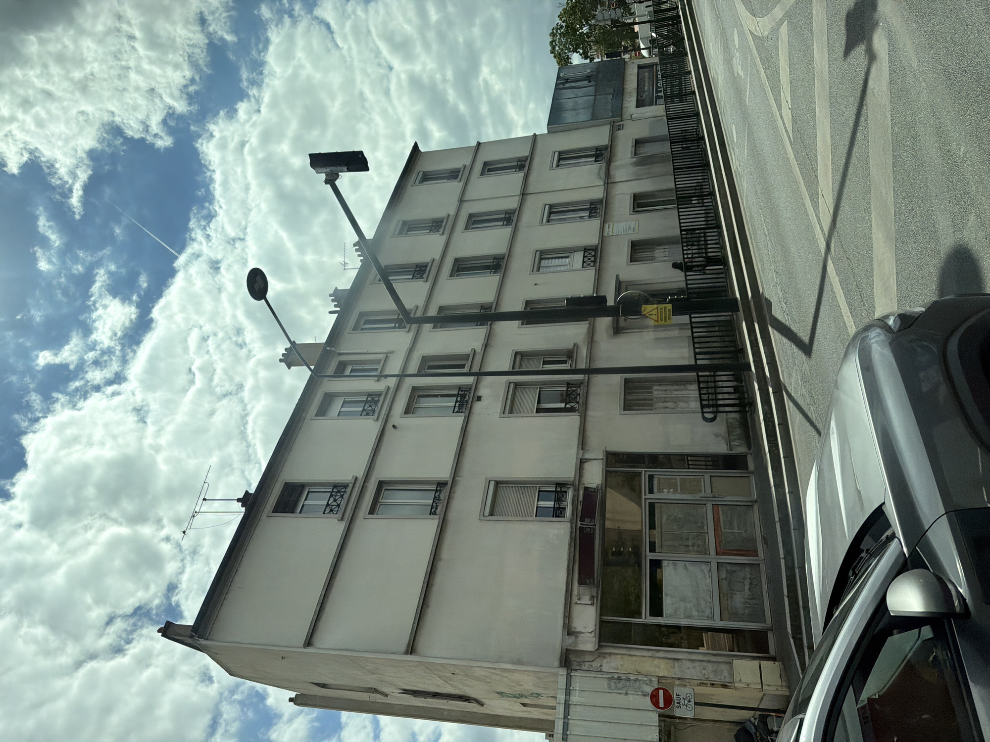 The image captures a street scene in Alfortville, France, focusing on a multi-story building and its surroundings. The building, with a pale facade, has several windows on each of its floors, some featuring decorative ironwork. The ground floor appears to be a commercial space with large glass windows, some of which are boarded up or obscured. A black metal railing runs along the front of the building at street level. A traffic light and a street lamp are prominently positioned in the foreground, casting shadows on the asphalt road. A gray car is partially visible in the lower left corner of the frame. The sky is filled with dramatic, white clouds against a blue sky, and a contrail from an airplane stretches across the upper right quadrant. A circular red and white sign with a bicycle symbol is affixed to a pole near the building's entrance. The overall impression is a somewhat overcast day in an urban environment.