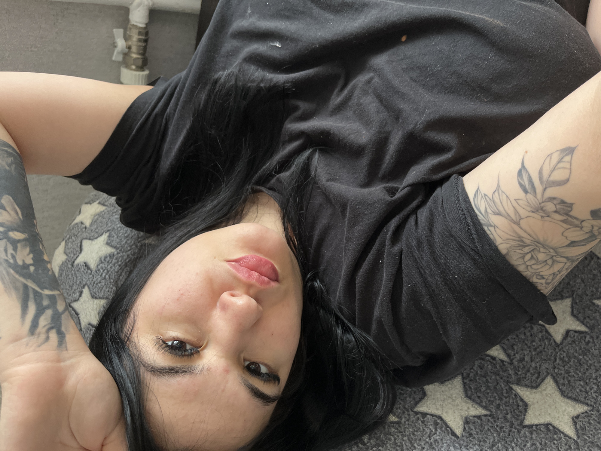A woman with dark hair lies on a star-patterned cushion. She wears a black t-shirt and has tattoos on her arms. A white pipe fixture is visible in the upper left corner. The location is Chaikovsky, Russia.