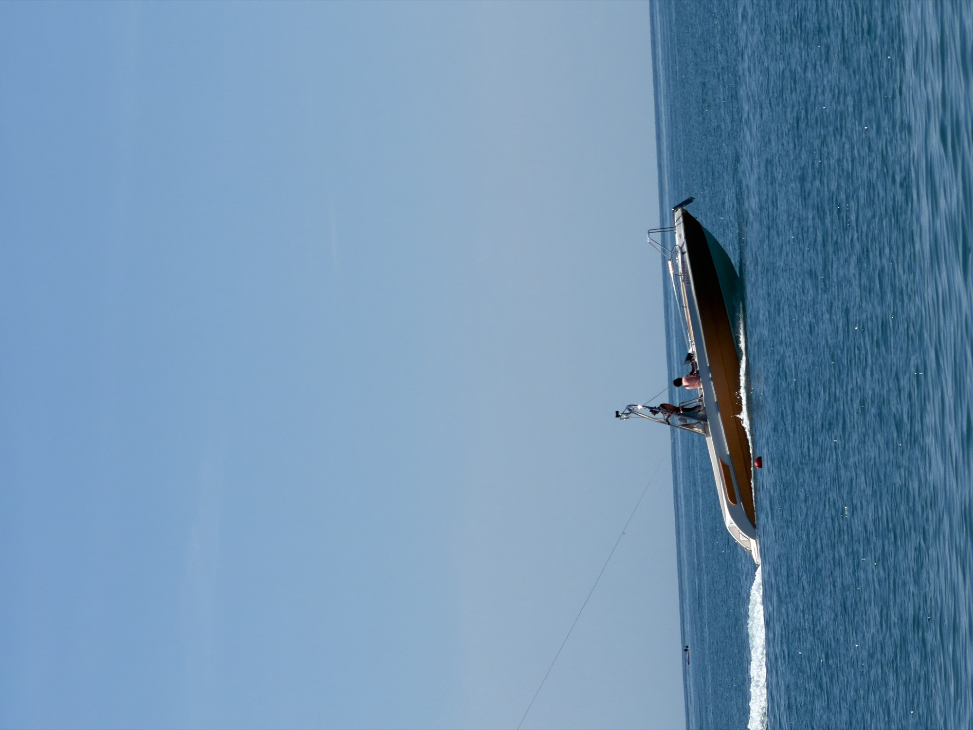 The image depicts a sailboat on the open sea, viewed from a high angle. The sky is a clear, pale blue, suggesting a sunny day. The sea is a deep blue, with whitecaps and a visible wake trailing the boat, indicating it is moving.
The sailboat itself is long and sleek, with a wooden deck and white hull. It appears to be a racing or performance sailboat. Two people are visible on board, one standing near the stern and another seated closer to the bow. They are dressed casually in t-shirts. The boat is angled diagonally in the frame, with its bow pointing towards the upper right corner of the image.
The water is textured with ripples and small white specks, possibly foam or sunlight reflecting on the surface. The wake of the boat is a prominent feature, a trail of white churned water against the blue. A thin rope or wire extends from the boat towards the left side of the frame, disappearing into the blue expanse.
The overall impression is one of a calm, perhaps leisurely sailing excursion on a clear day. The scene is devoid of any urban or land-based elements, emphasizing the vastness of the ocean and sky. There is no visible text in the image. The orientation of the image suggests it may have been taken from another boat or a drone.