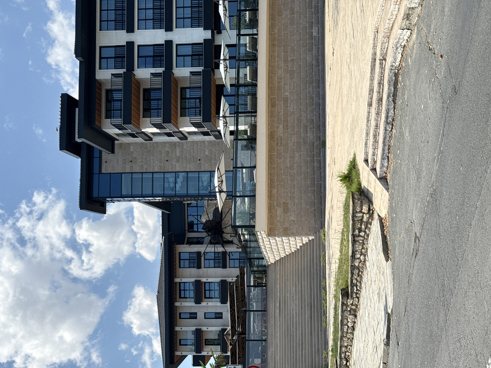 The image shows a modern hotel building with multiple stories, featuring white facades, dark balconies, and large windows. The hotel has a distinctive glass-enclosed section. In front of the hotel, there is a paved area with lounge chairs and umbrellas, indicating an outdoor seating or relaxation area. A wide set of concrete stairs leads up to this area from a lower level. The building is constructed with stone and has a flat, dark roof. The sky is blue with scattered white clouds. The surrounding environment appears to be paved, with some patches of grass and a low stone wall bordering the paved area. The scene suggests a sunny day, possibly in a resort or coastal town. This hotel is located in Nesebar, Bulgaria.