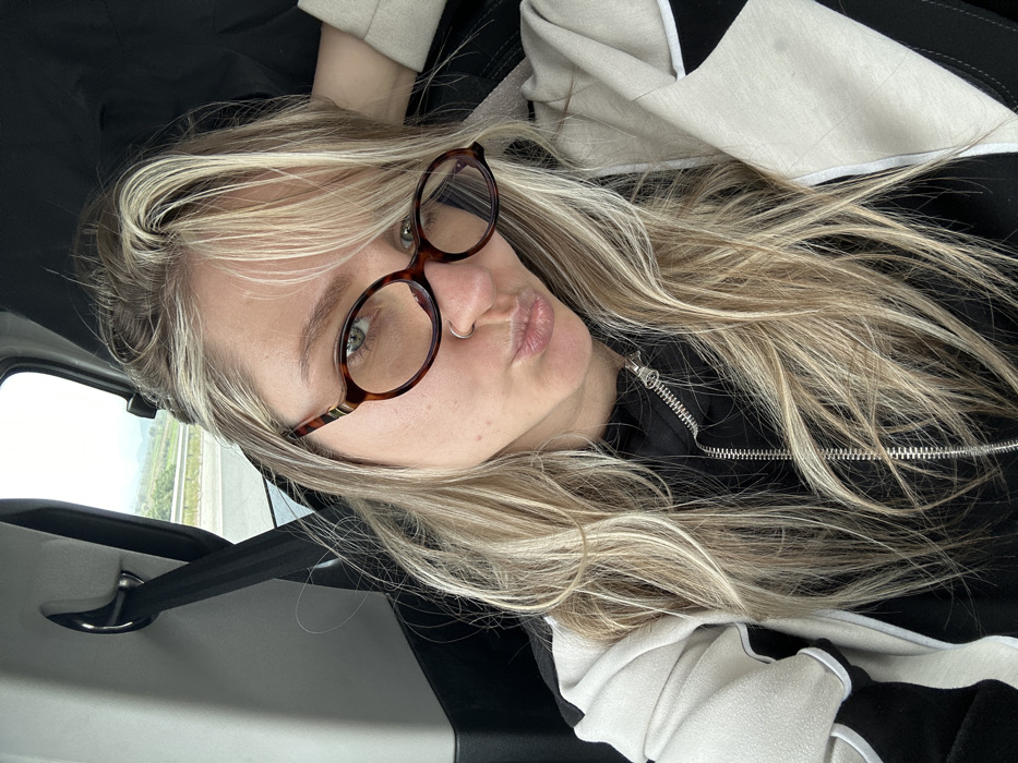 A female subject with long blonde hair, dark roots, green eyes, and a small silver ring in her left nostril is depicted in a close-up, seated within a vehicle. She wears tortoise-shell eyeglasses and a light-colored jacket with black and white accents over a black zippered garment. Her lips are pursed forward. Her left hand is raised, partially obscured, with fingers resting behind her head. To the subject's left, a black vehicle seatbelt is visible. Through the vehicle window, a multi-lane road or highway is visible, running alongside a landscape of green hills and sparse vegetation under an overcast sky. This environmental setting is consistent with the rural and semi-urban landscapes found along major roads in the Torbalı district of İzmir Province, Turkey.