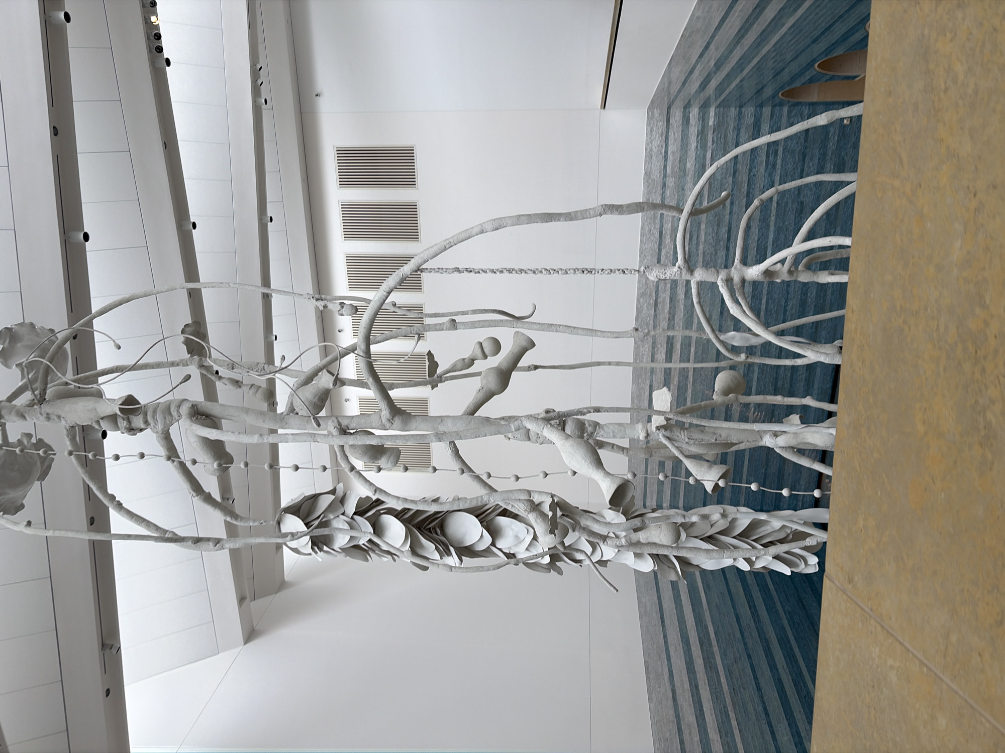 This image depicts a white art installation hanging in an indoor space. The installation features branch-like structures with various sculptural forms attached, resembling leaves, vessels, and abstract shapes. It is suspended between white walls and a carpeted floor with blue and gray stripes. The background includes a ventilation grill on the wall and architectural details such as beams with mounted lights. A tan-colored wall is visible on the right. This art installation is located in the lobby of the Austin Proper Hotel in downtown Austin, United States of America.