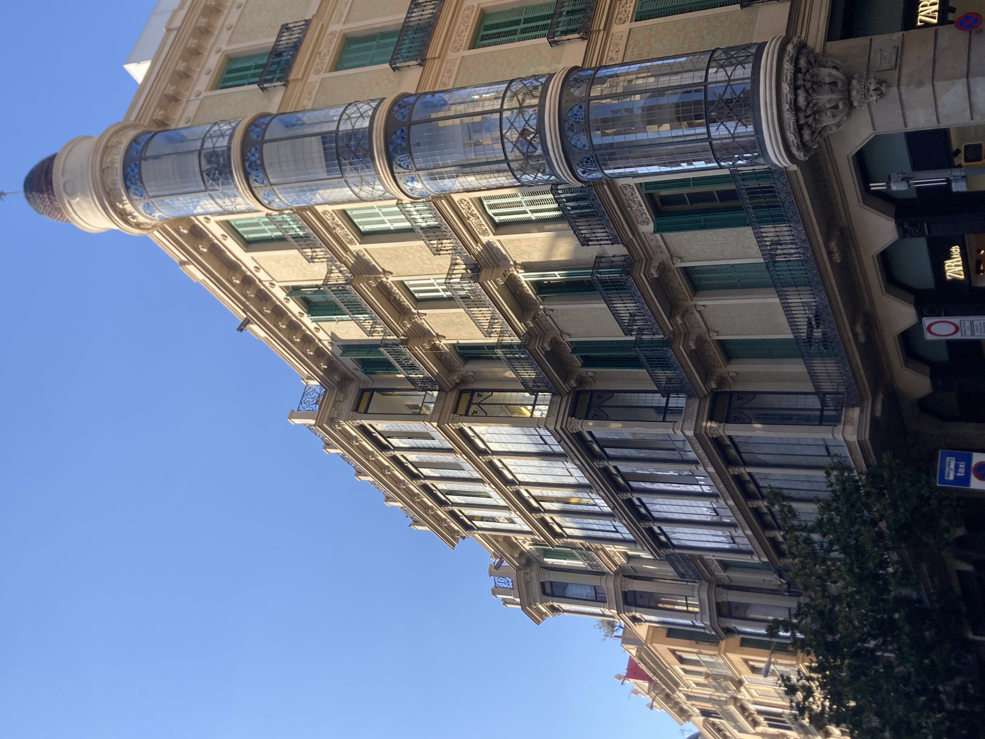 This is a low-angle, eye-level, outdoor, daytime, full shot of the facade of a historic building in the Eixample district of Barcelona, Spain. The building, with its distinctive curved corner featuring a glass tower and ornate decorative elements, is bathed in sunlight. The facade is a light beige color with green shutters on many of the windows. Balconies with black wrought-iron railings extend from the windows. Below, the ground floor appears to house retail establishments, with signage for "ZARA" and "ZARA Kids" visible. A tree partially obscures the lower left portion of the building. The sky above is clear and blue.