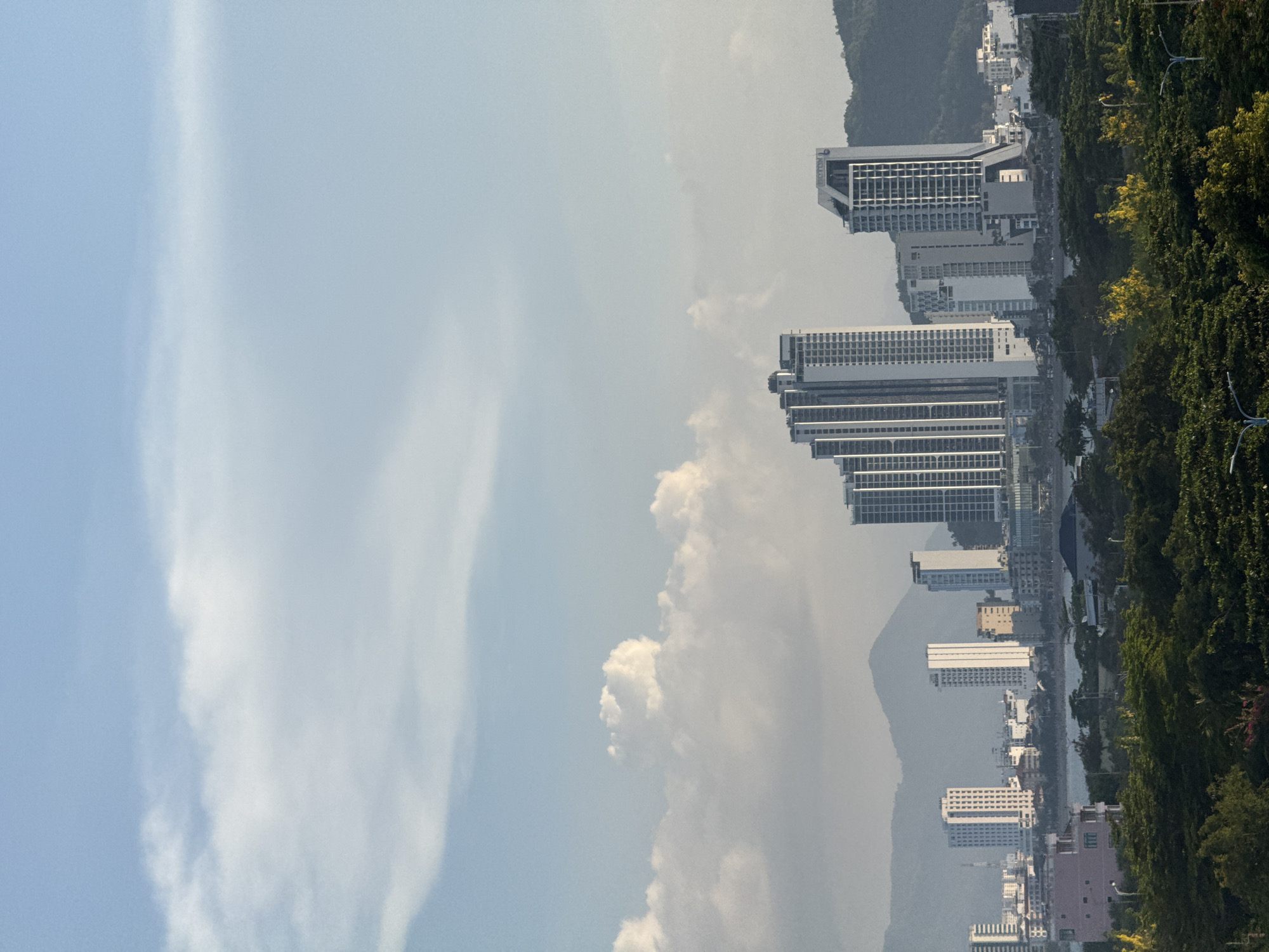 A wide shot captures a coastal city skyline in Bac Nha Trang Ward, Vietnam, under a bright, partially clouded sky. The foreground is dominated by a dense line of lush green trees. Behind the trees, the city unfolds with numerous modern high-rise buildings, many appearing to be residential or commercial towers, clustered along the waterfront. Mountains form a hazy backdrop to the urban landscape. A body of water, likely the sea, is visible between the trees and the city. The overall impression is of a vibrant, developing coastal city.