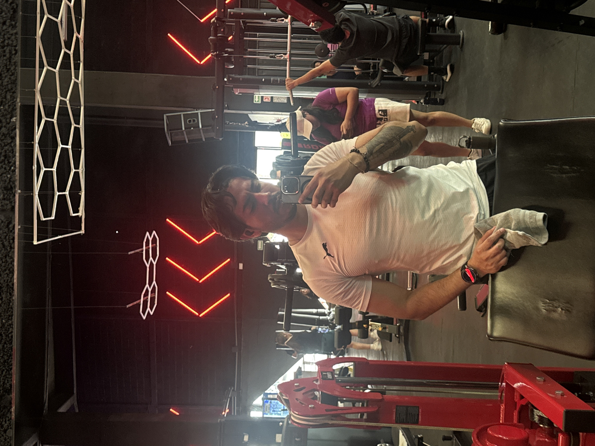 A man wearing a white t-shirt is taking a selfie in a gym. The gym is located in Puebla, Mexico. In the background, other people are working out on various weight machines. The gym has dark walls with geometric neon lighting fixtures. The man is holding a cell phone in his right hand, and a towel is visible on the seat behind him. He is wearing a wristwatch on his left wrist. Red gym equipment is in the foreground.
