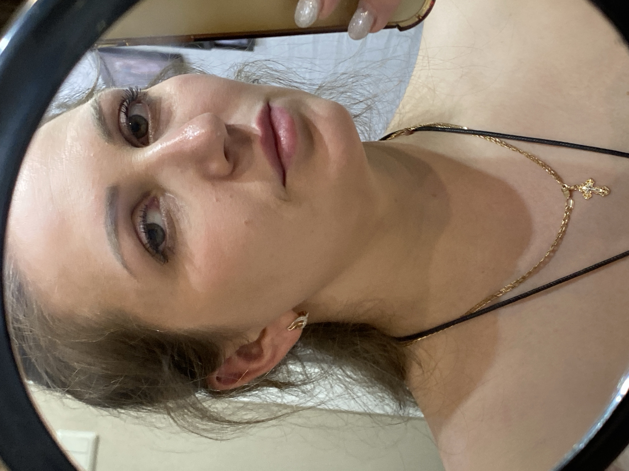 A close-up, tilted, selfie-style view shows a young woman reflected in a handheld mirror. The woman has fair skin, brown hair, and light brown eyes. She is wearing a gold chain necklace with a cross pendant and small gold earrings. The background, visible through the mirror, appears to be a room with light-colored walls and indistinct furniture. A hand with glittery nails holds the mirror. There is no indication of Markovskii, Russia.